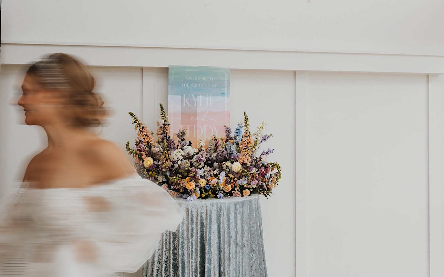 A bride runs across the photo, behind her is a table with flowers on it and a custom sign for her wedding at The Aisling