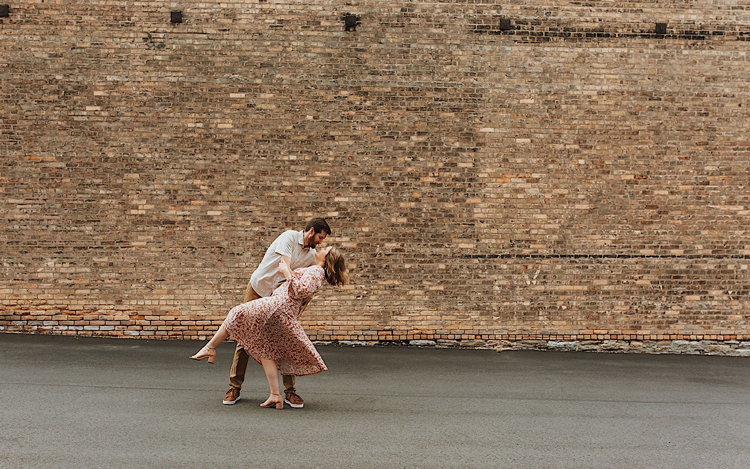 While taking engagement photos in downtown Minneapolis a man dips a woman as the two dance in front of a brick wall