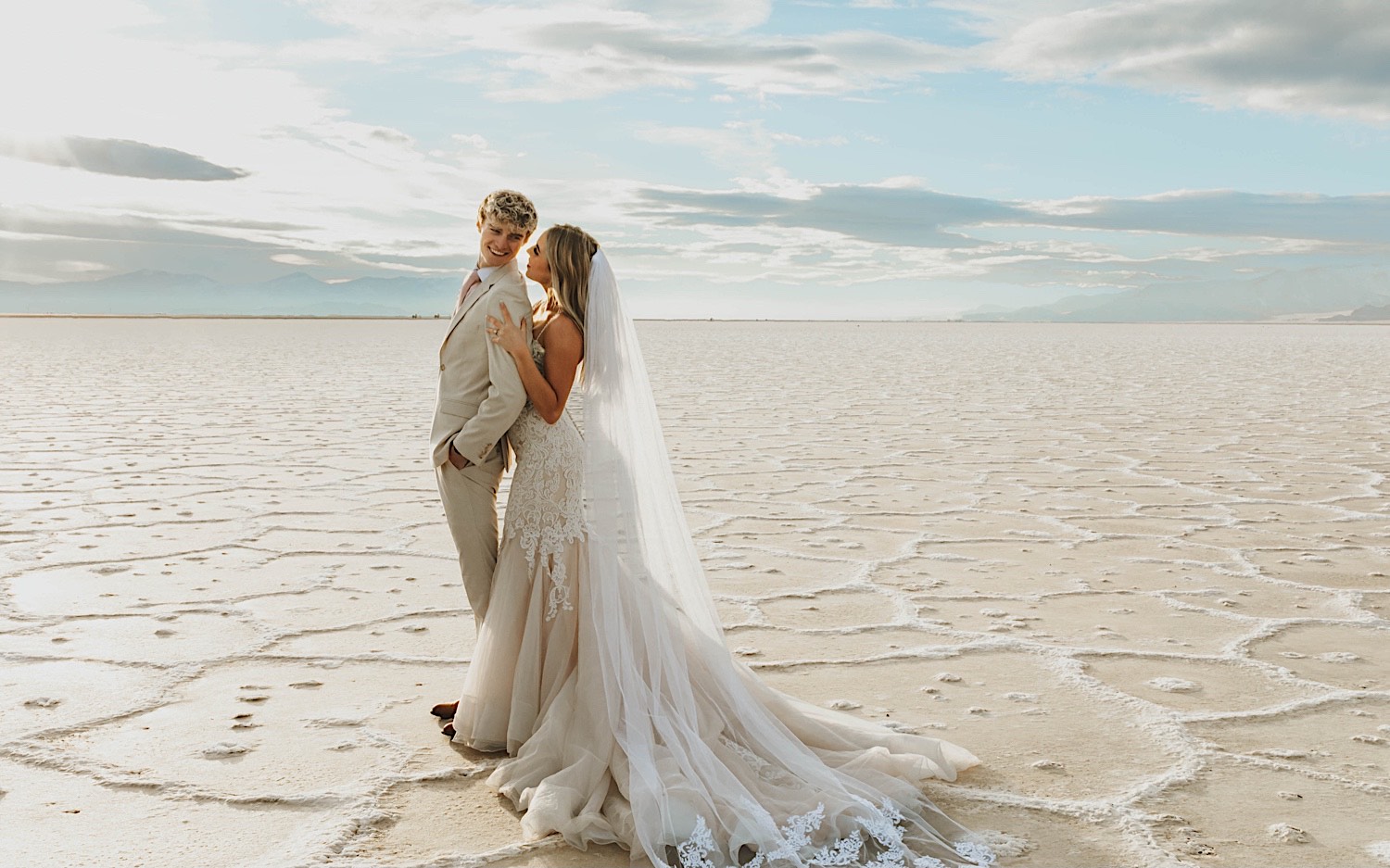 A groom smiles over his shoulder as the bride stands behind him and touches his shoulder as the two stand in the Utah Salt Flats on their elopement day