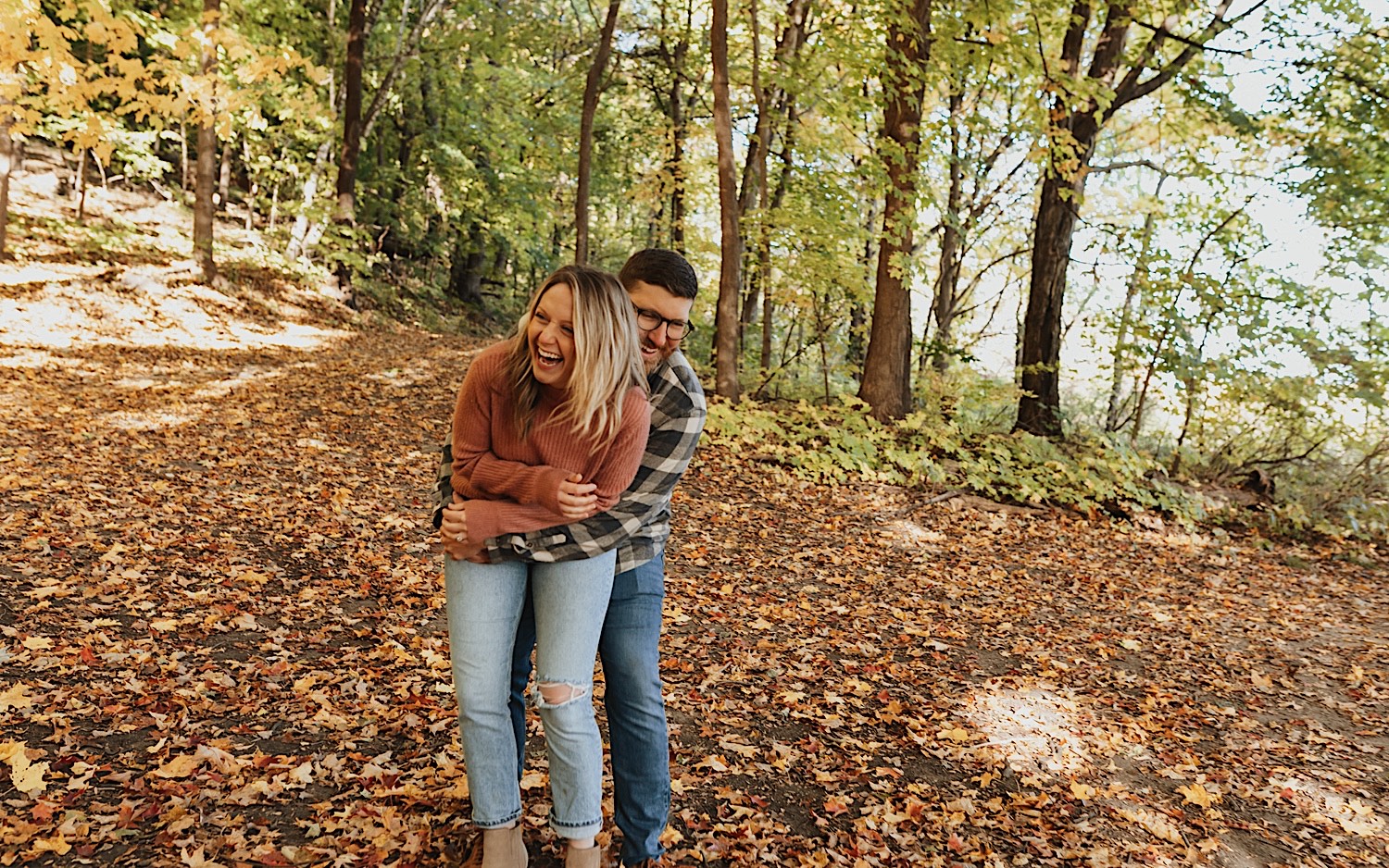 During their fall engagement session in Winona a couple laugh and smile while standing on a leaf covered trail in the forest as the man hugs the woman from behind