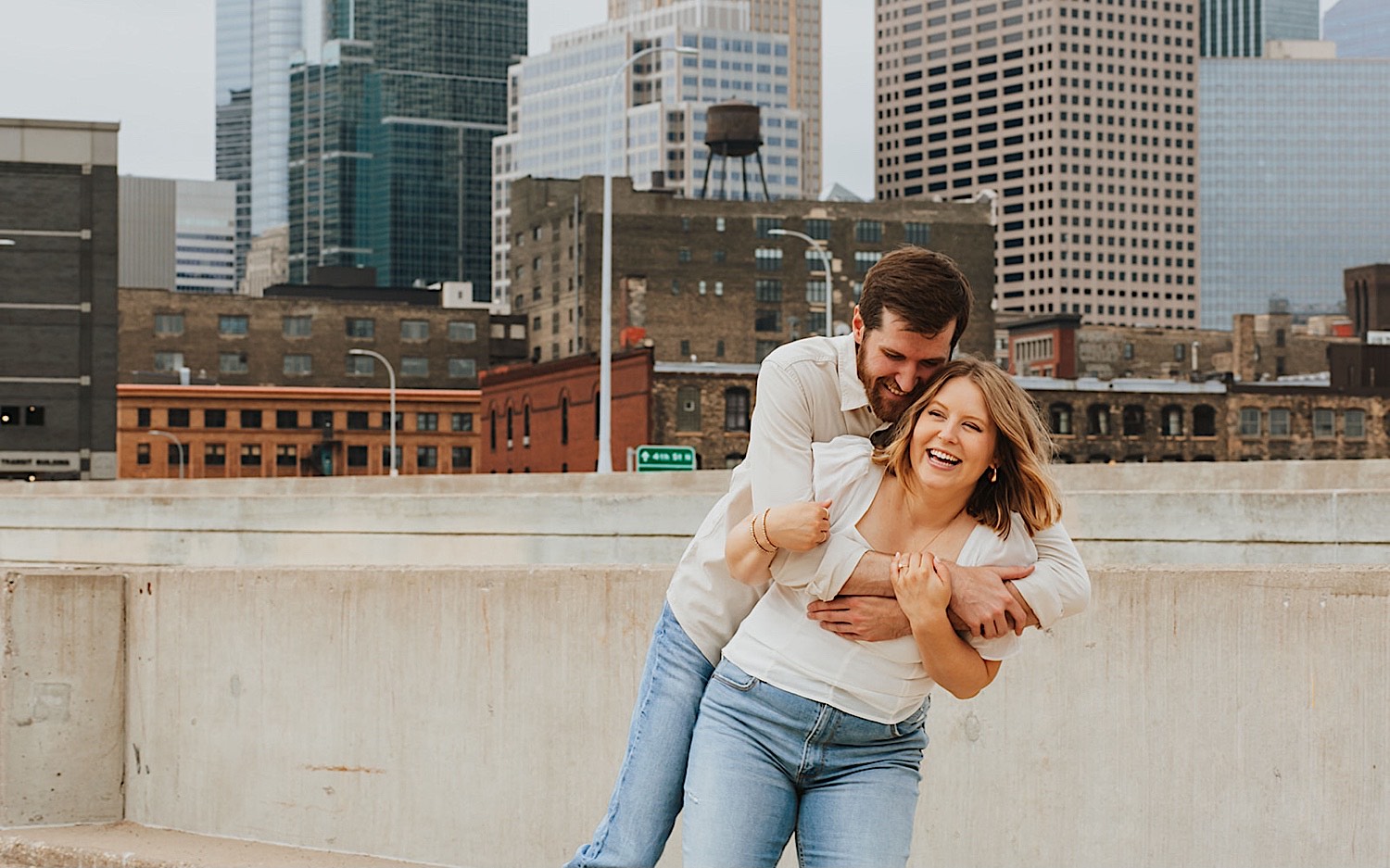 While on the top of a parking garage in downtown Minneapolis a man hugs a woman from behind as she smiles while they take their engagement photos