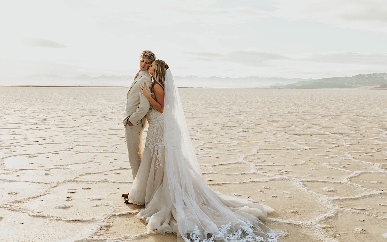 A groom looks over his shoulder as the bride embraces him from behind and kisses him on the shoulder, they're standing in the Utah Salt Flats on the day of their elopement