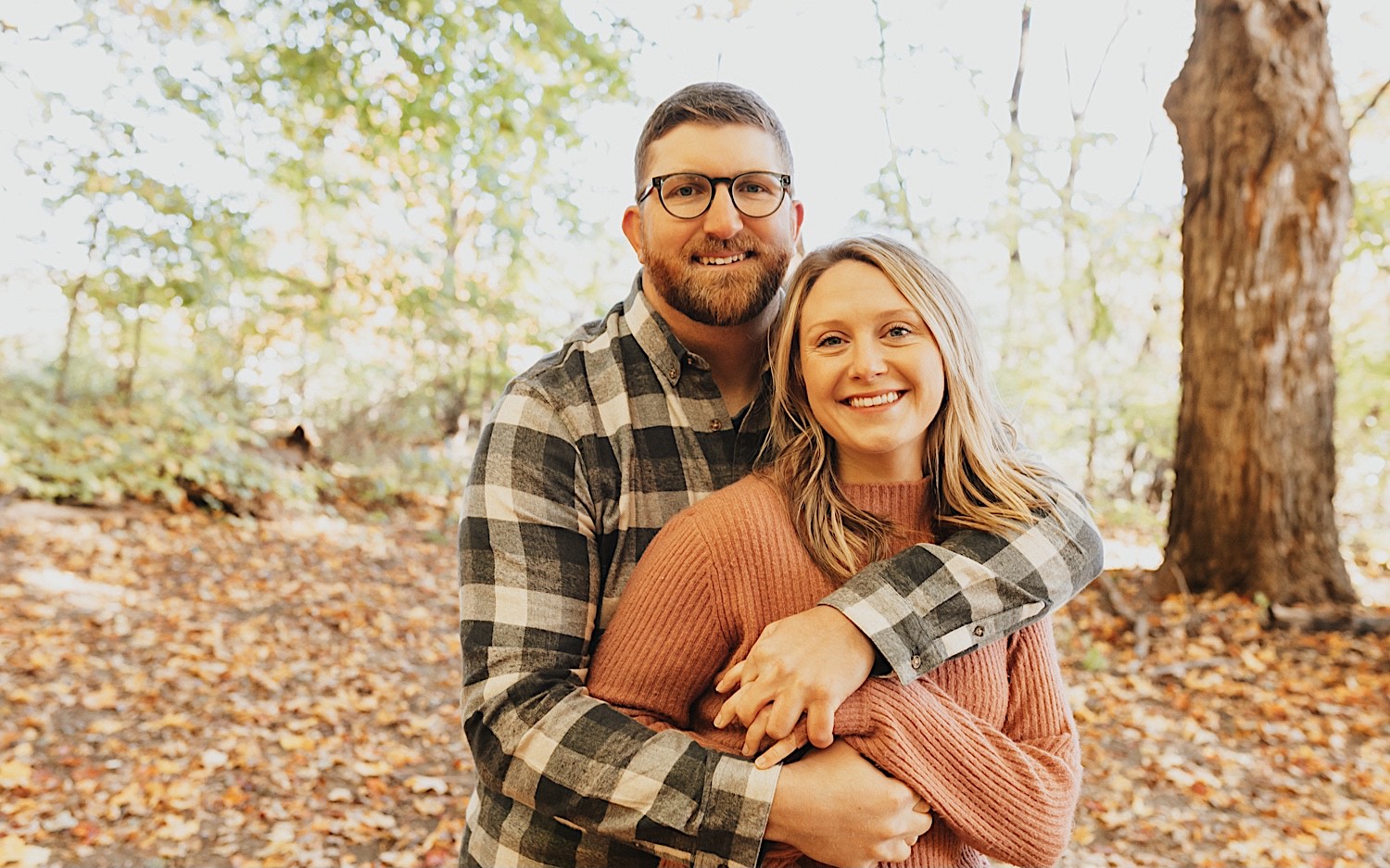 Portrait photo of a man and woman where the man is hugging her from behind and they are both smiling at the camera with leaves on the ground around them
