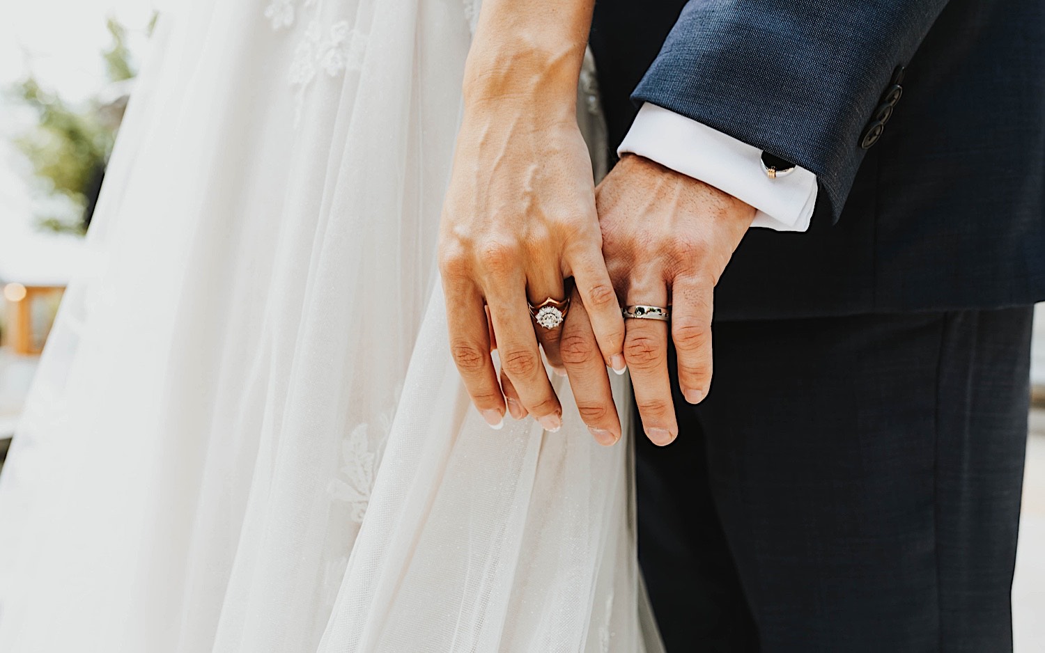 Close up photo of a bride and groom's hands holding one another, each with their wedding ring being shown off to the camera