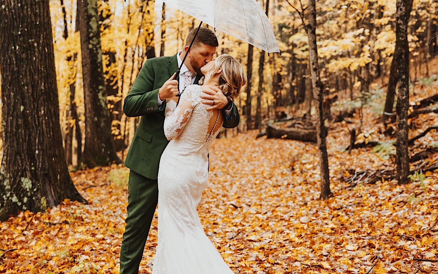A bride and groom kiss one another while under an umbrella in the middle of a forest covered in yellow leaves during their wedding at Lake Bomoseen Lodge in Vermont