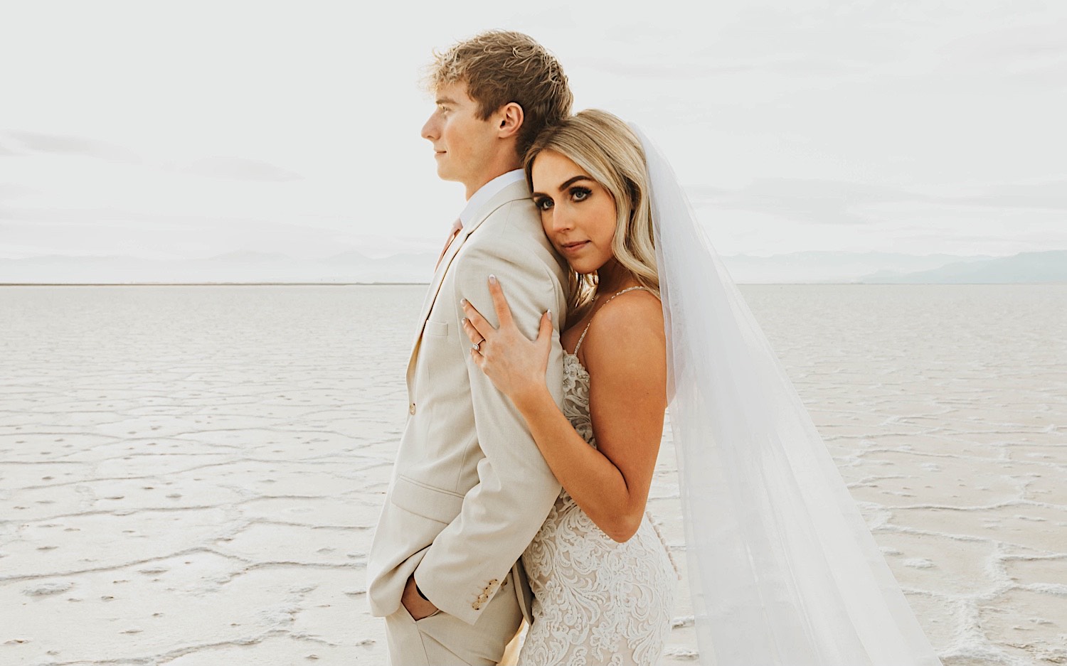 A groom looks off camera as the bride embraces him from behind and looks towards the camera while standing in the Utah Salt Flats on the day of their elopement