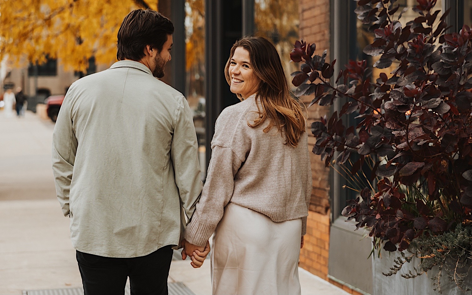 A woman smiles back over her shoulder while walking hand in hand with a man during their engagement session in Minneapolis' North Loop