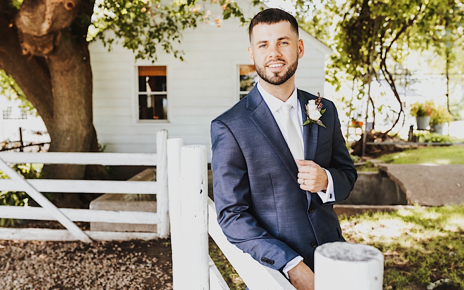 A groom smiles at the camera while leaning against a fence at the wedding venue Legacy Hill Farm