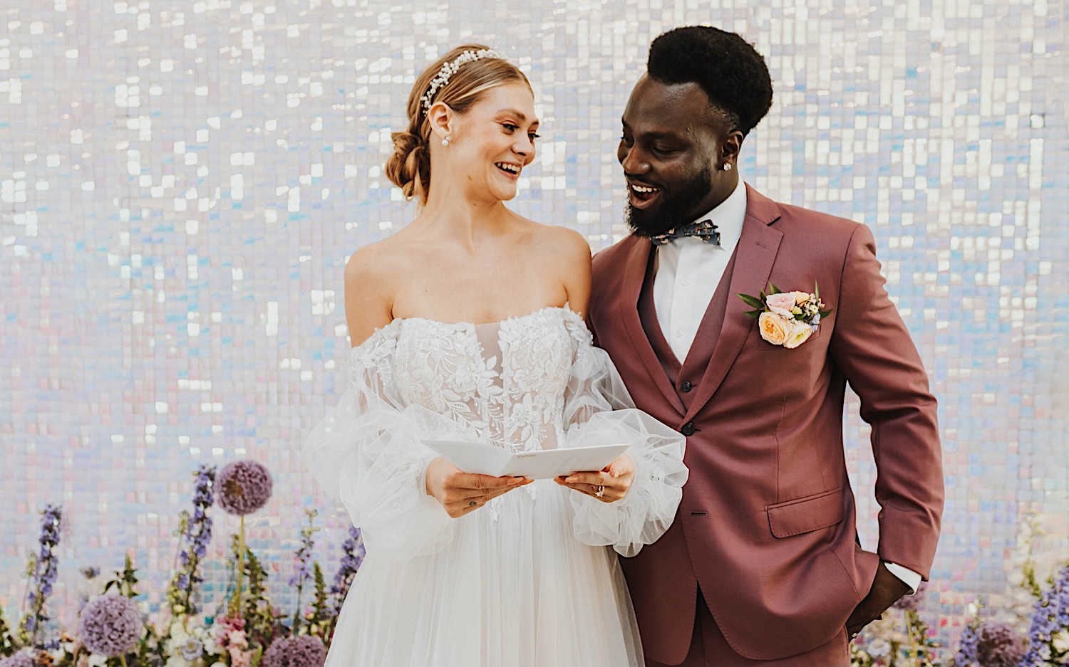 A bride and groom standing in front of a sparkling wall smile as they read a letter together during their wedding day at The Aisling