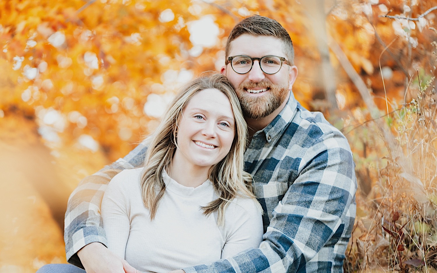 A couple sit with one another and smile at the camera in front of fall leaves on a tree during their engagement session in Minnesota A couple sit with one another and smile at the camera in front of fall leaves on a tree during their engagement session in Minnesota