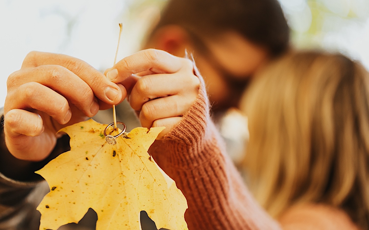 Detail photo taken during a fall engagement session in Winona of a yellow leaf being held by a couple who are kissing one another in the background, on the leaf's stem is an engagement ring