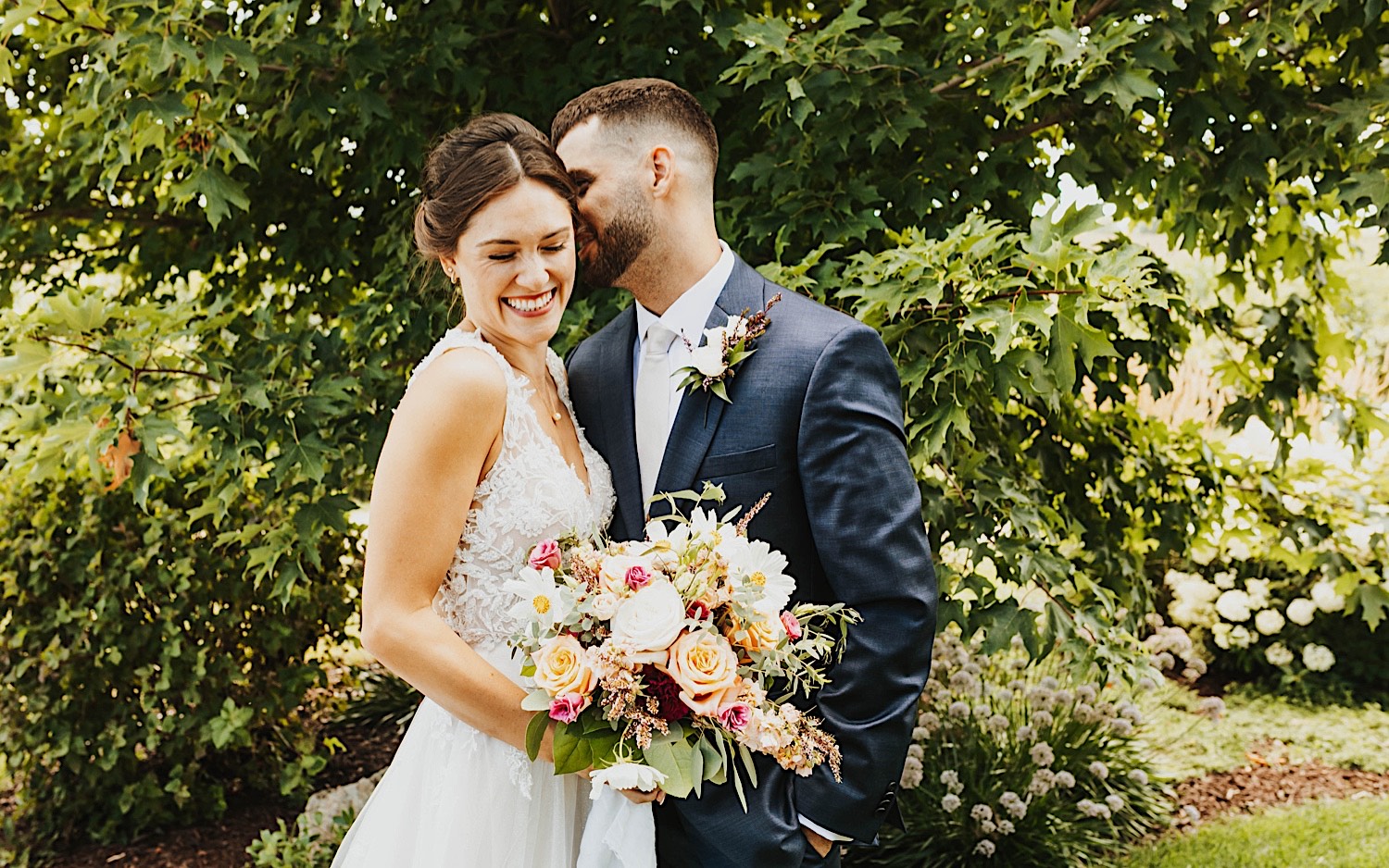 A bride smiles as the groom whispers in her ear while the two stand in front of a tree on the grounds of their wedding venue Legacy Hill Farm