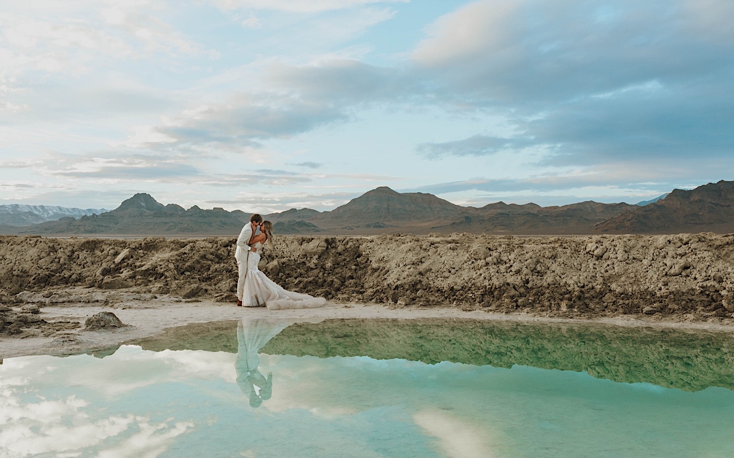 During their elopement in the Utah Salt Flats a bride and groom stand behind a pool of water in the mountain range and embrace as they're about to kiss one another