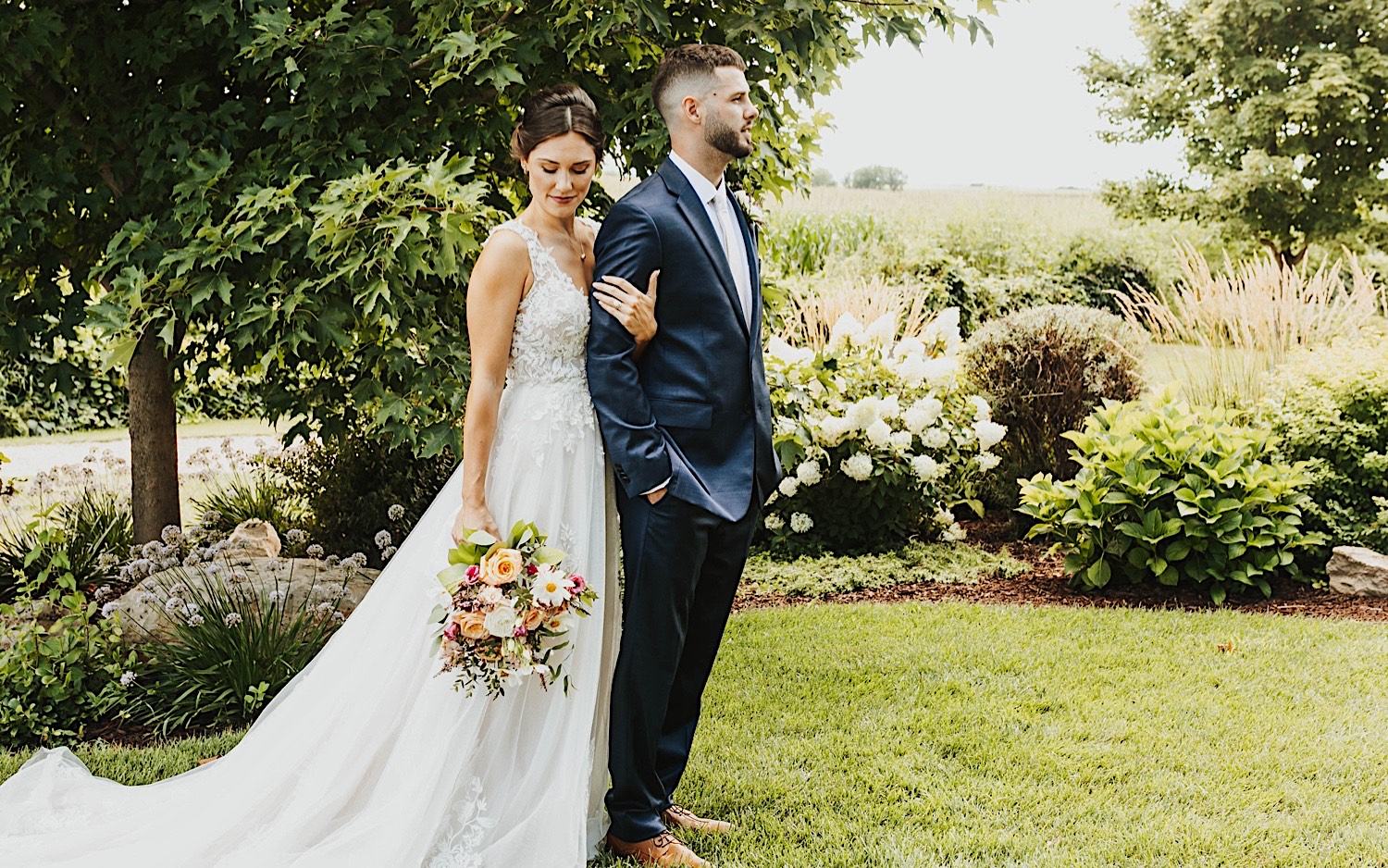 A bride stands behind the groom and locks arms with him while they stand outdoors at their wedding venue Legacy Hill Farm A bride stands behind the groom and locks arms with him while they stand outdoors at their wedding venue Legacy Hill Farm