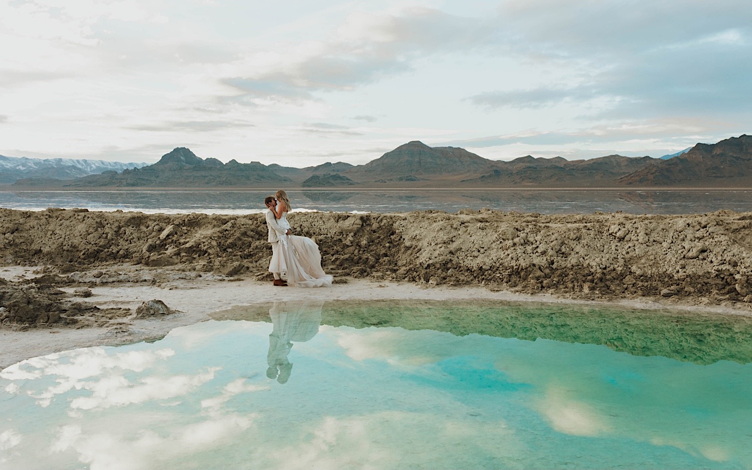 In the Utah Salt Flats, a bride is lifted in the air by the groom with a pool of water in front of them on their elopement day