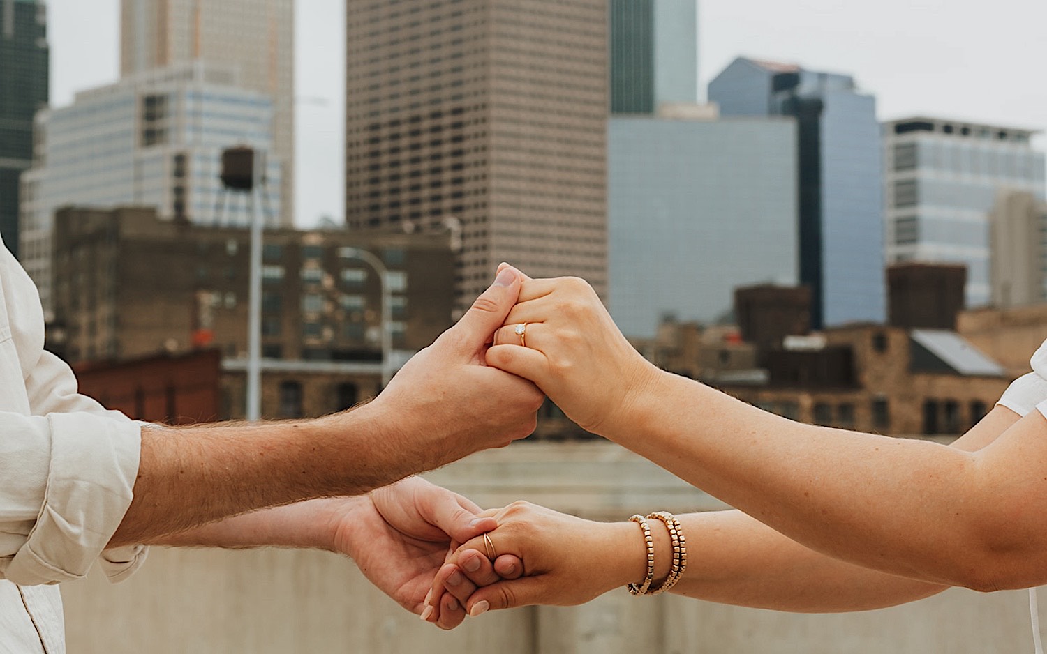 Close up photo of a couple's hands holding one another showing off the engagement ring on the woman's hand, behind them is the skyline of downtown Minneapolis