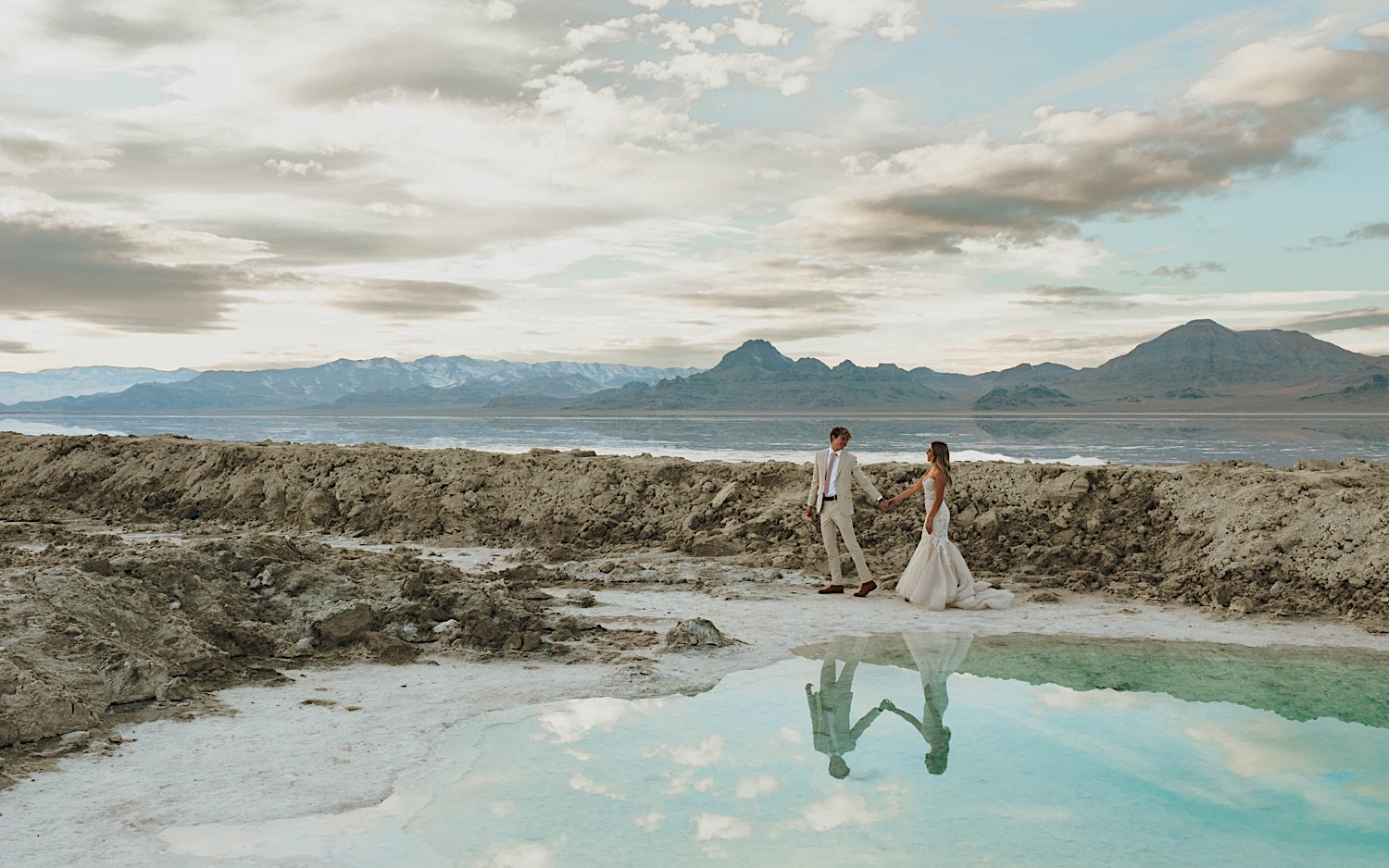 During their elopement in the Utah Salt Flats, a groom leads the bride by her hand and smiles back at her as they walk next to a pool of water with mountains in the background