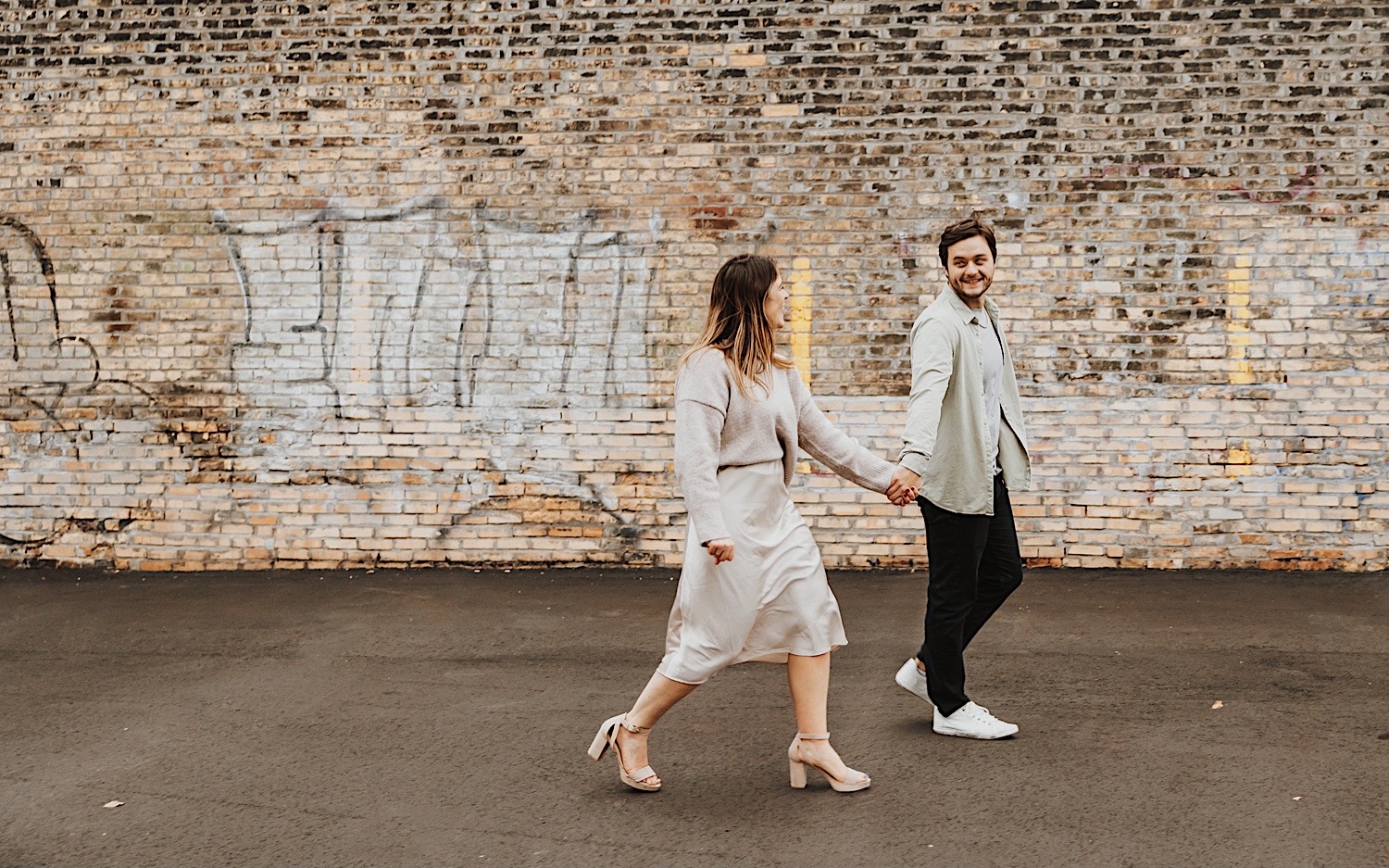 A couple smile at one another while walking hand in hand in front of a brick wall in Minneapolis' North Loop during their engagement session