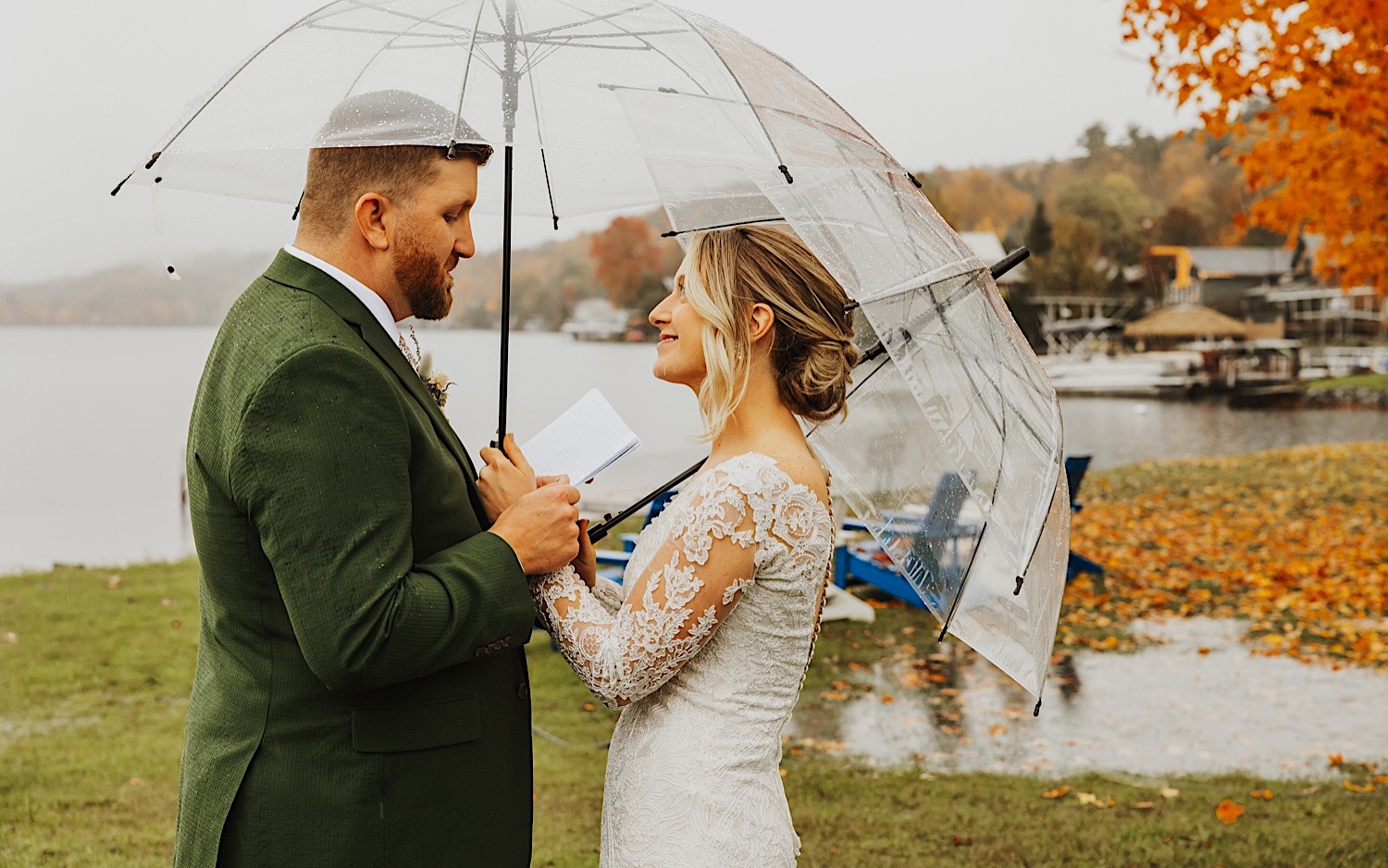 A bride smiles while the groom reads his private vows to her while the two stand under clear umbrellas next to Lake Bomoseen in Vermont