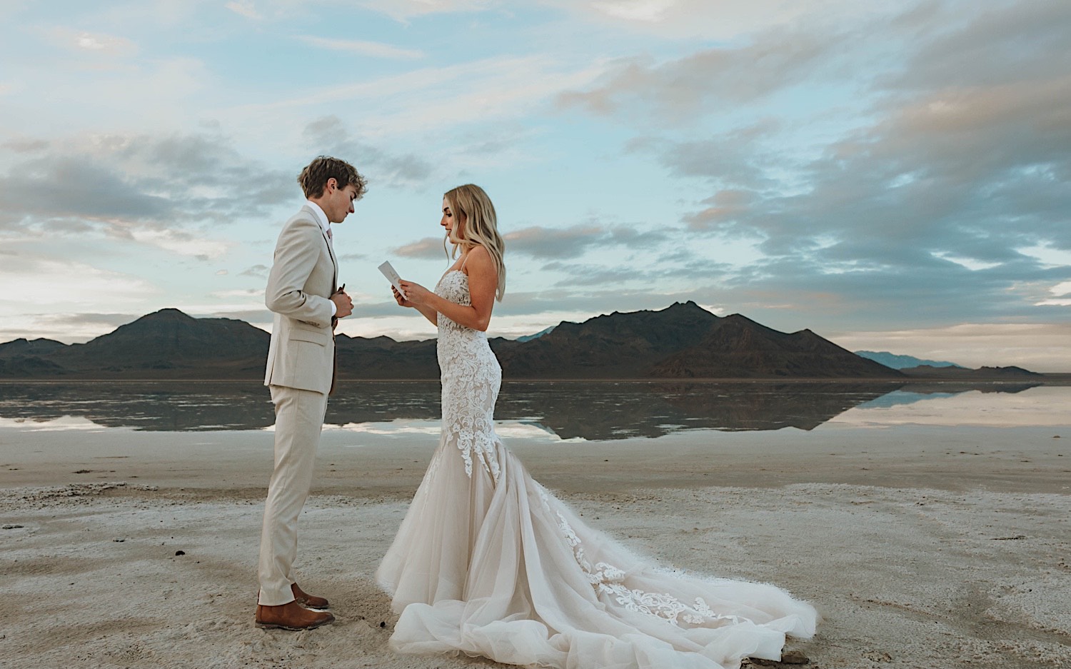 During their elopement in the Utah Salt Flats, a bride and groom stand facing one another and read their vows to each other with mountains behind them