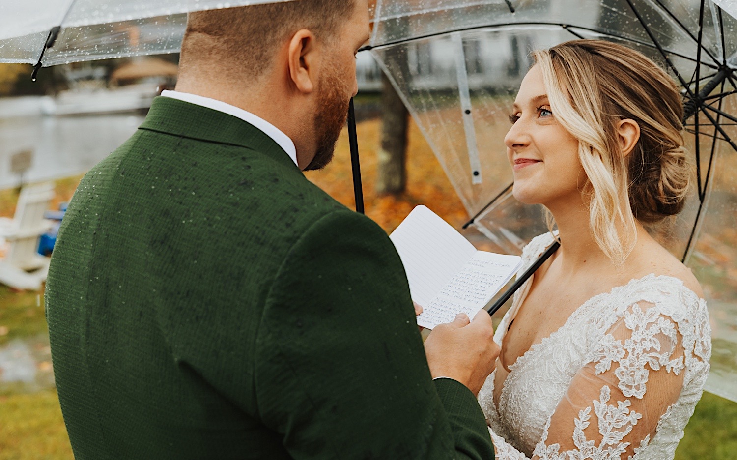 A bride looks to the groom as he reads his vows to her while the two stand under umbrellas on a rainy day at Lake Bomoseen in Vermont