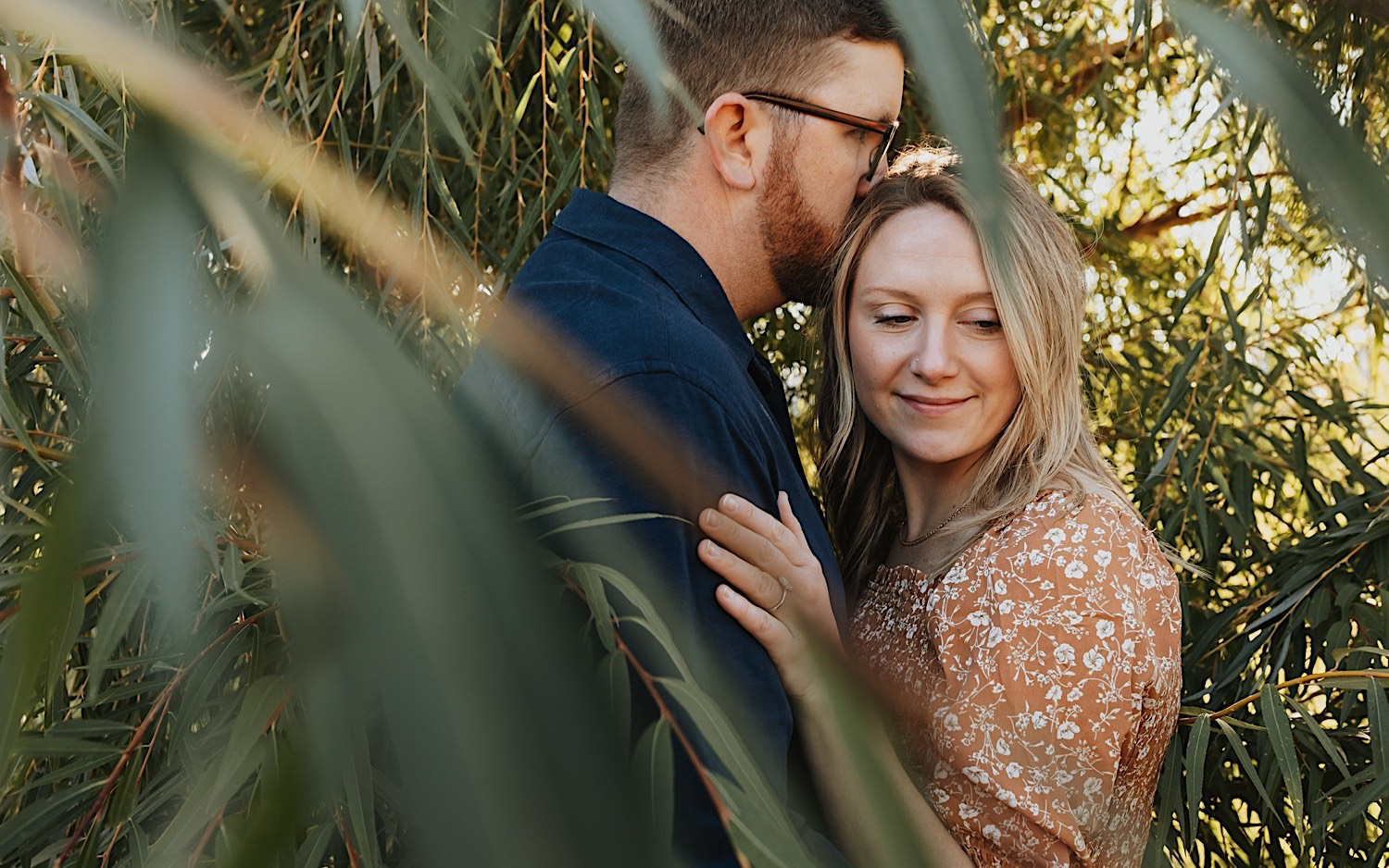 During a fall engagement session in Winona a couple embrace as the man kisses the woman on the temple while she smiles and looks to the right as the two are surrounded by the leaves of a tree