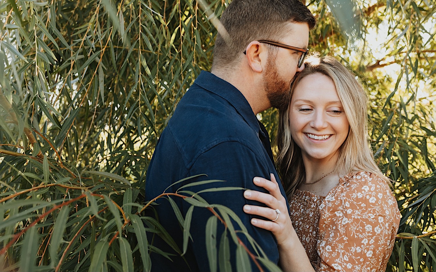 During a fall engagement session in Winona a couple embrace as the man kisses the woman on the temple while she smiles and looks to the right as the two are surrounded by the leaves of a tree