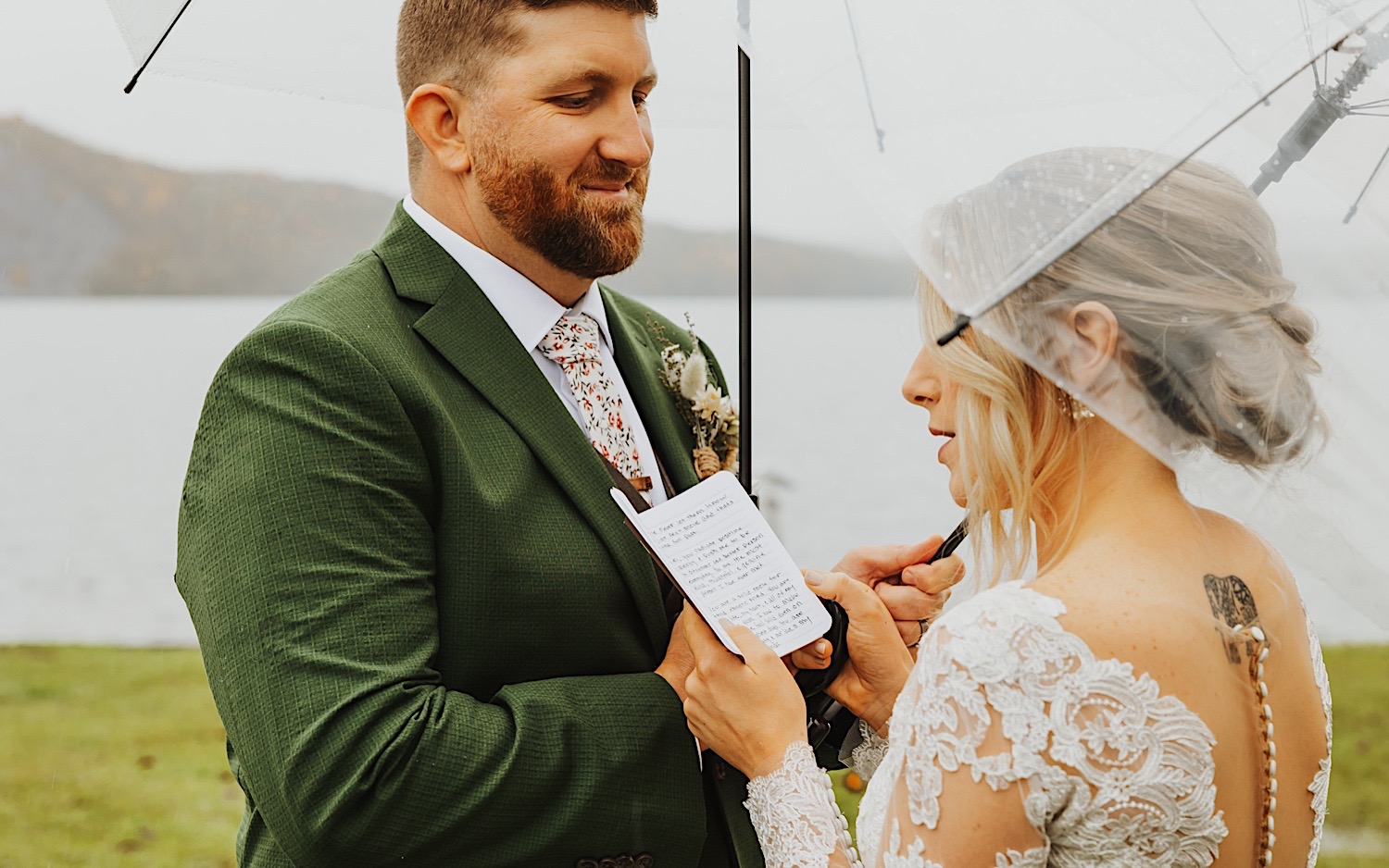 A groom smiles as the bride reads her vows to him while the two stand under umbrellas on a rainy day in Vermont next to Lake Bomoseen