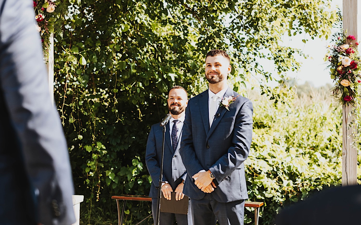 A groom stands and smiles with hands clasped while at the outdoor ceremony space of the wedding venue Legacy Hill Farm A groom stands and smiles with hands clasped while at the outdoor ceremony space of the wedding venue Legacy Hill Farm