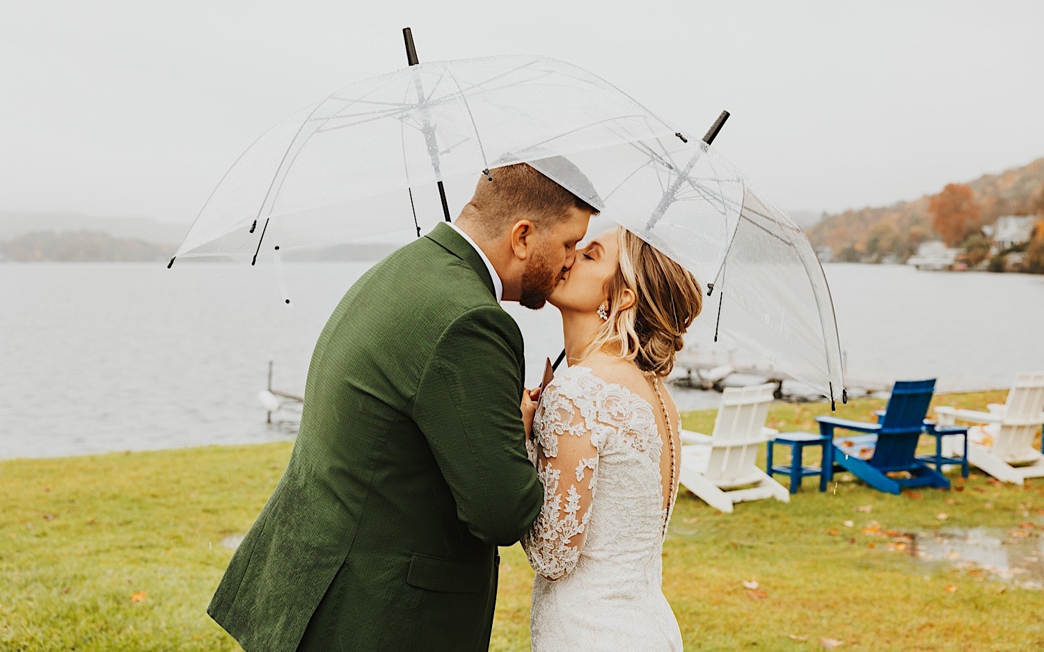 A bride and groom kiss while under clear umbrellas as they stand next to Lake Bomoseen in Vermont