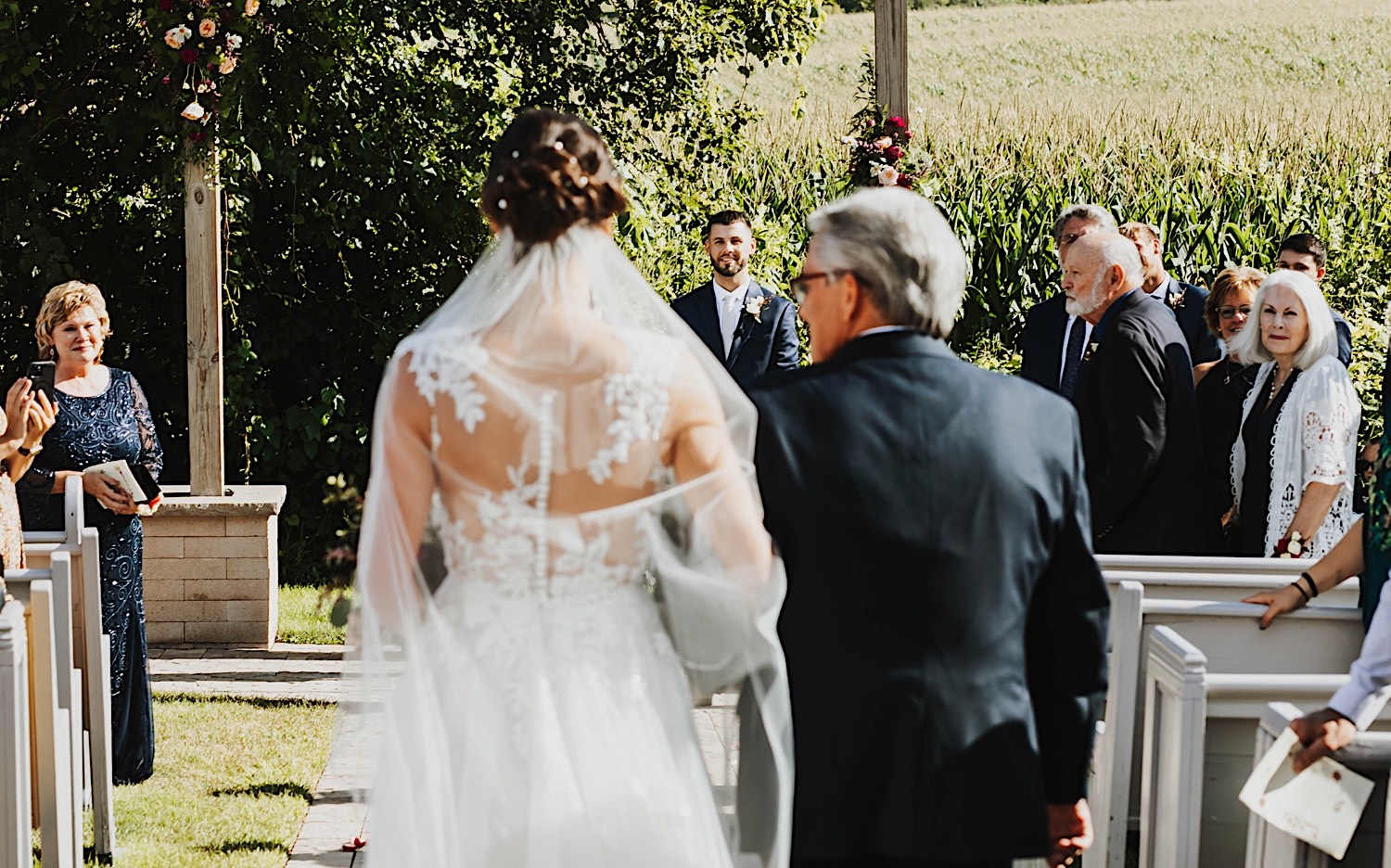 A groom smiles in the background as the bride and her father walk down the aisle together in the foreground of the photo during the outdoor wedding ceremony at Legacy Hill Farm