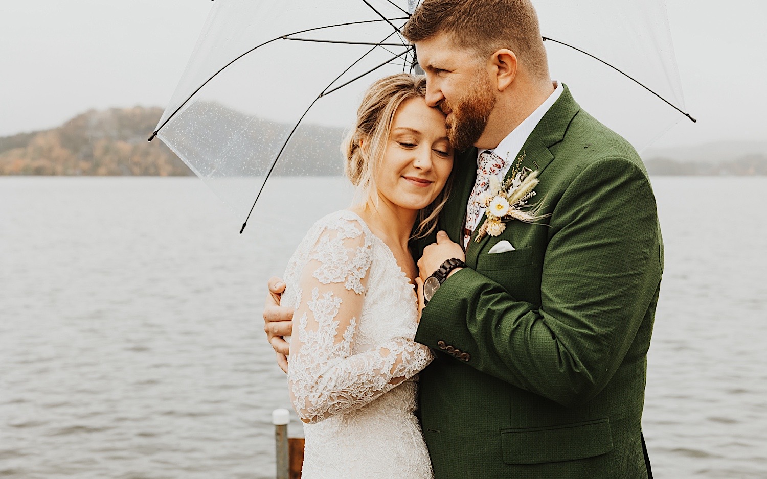 A bride smiles and closes her eyes as the groom hugs her and kisses her forehead while the two stand under an umbrella on a dock of Lake Bomoseen in Vermont