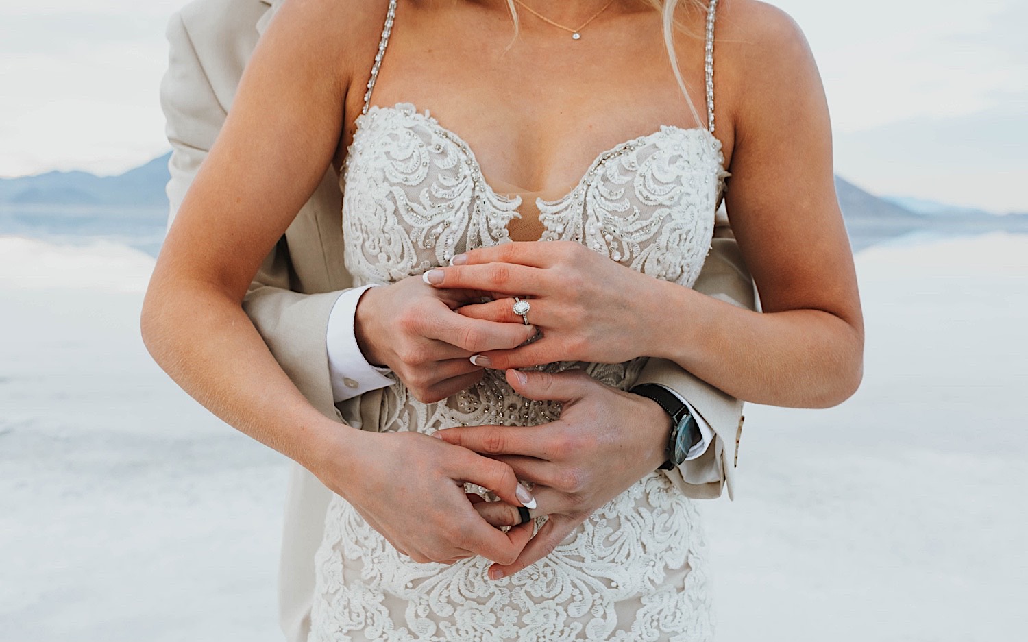 Close up photo of a bride and grooms hands as the groom embraces the bride from behind, the photo shows off their wedding rings during their elopement in the Utah Salt Flats