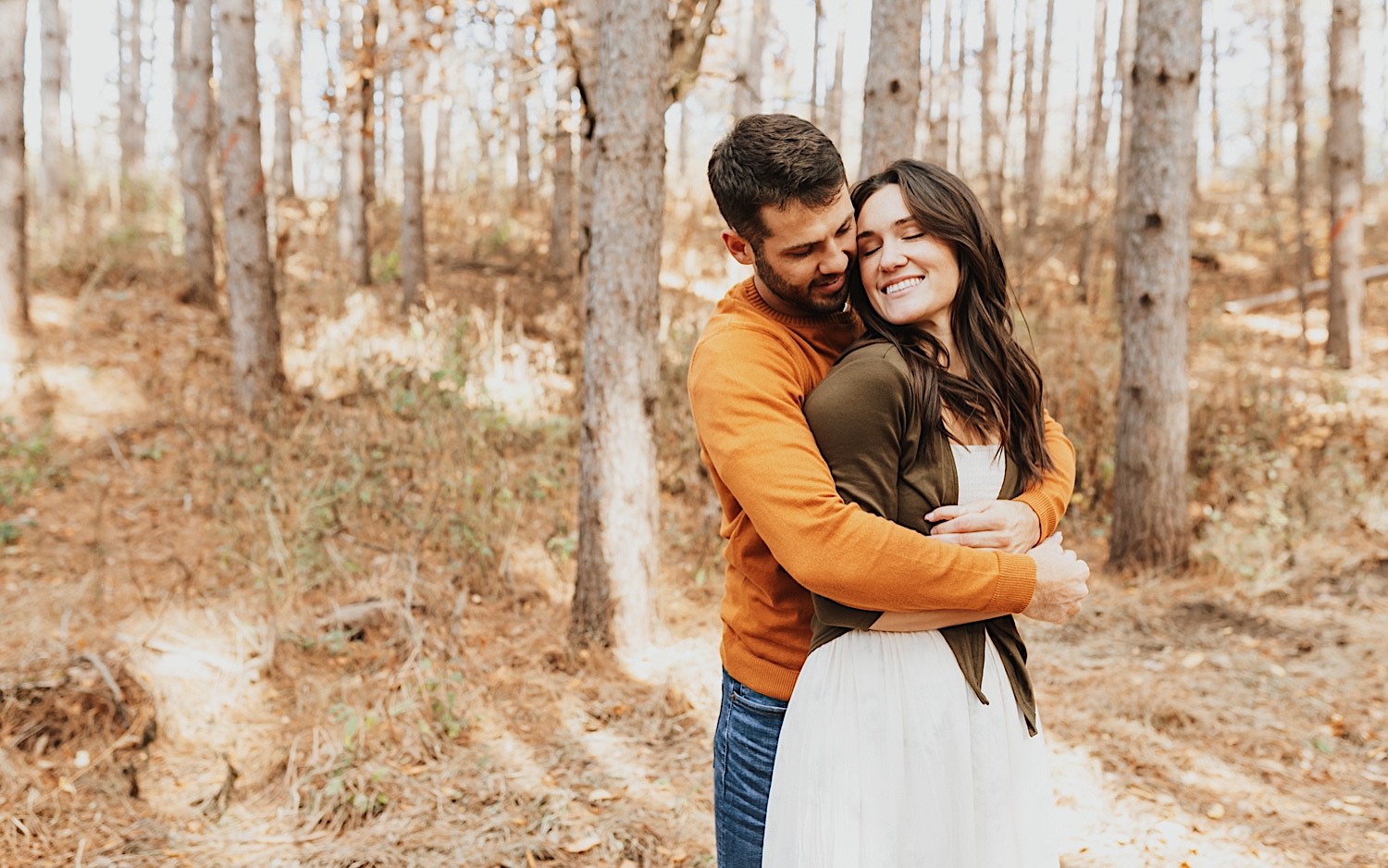 A woman smiles with her eyes closed as a man hugs her from behind while they stand in a forest with fall leaves on the ground during their engagement session in Minnesota A woman smiles with her eyes closed as a man hugs her from behind while they stand in a forest with fall leaves on the ground during their engagement session in Minnesota
