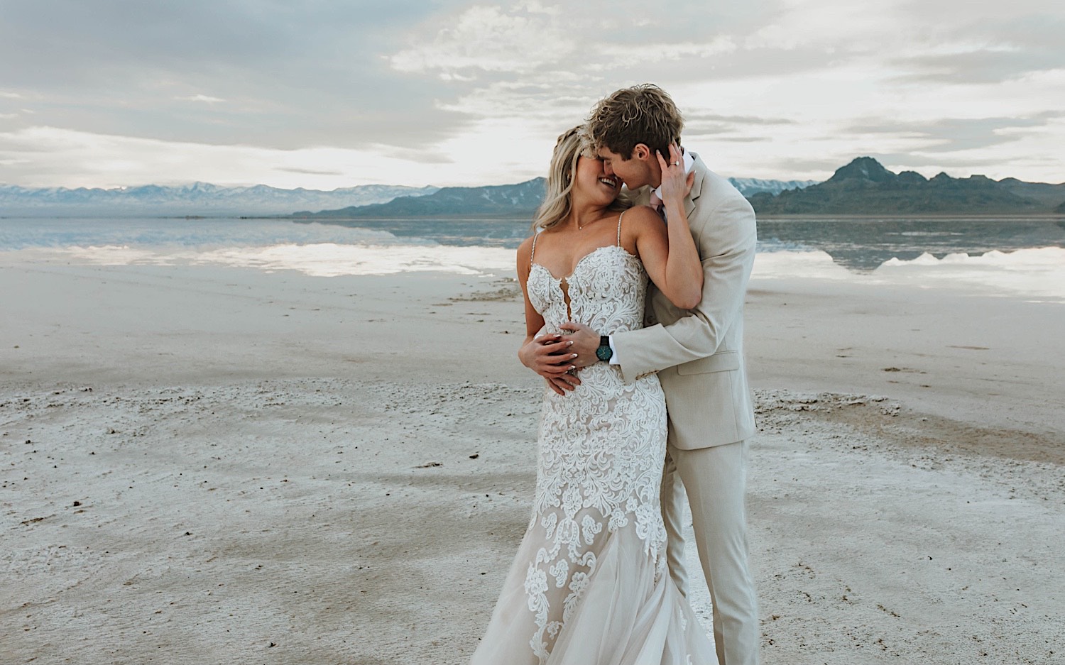 In the Utah Salt Flats a groom embraces the bride from behind during their elopement and are about to kiss one another as the bride smiles