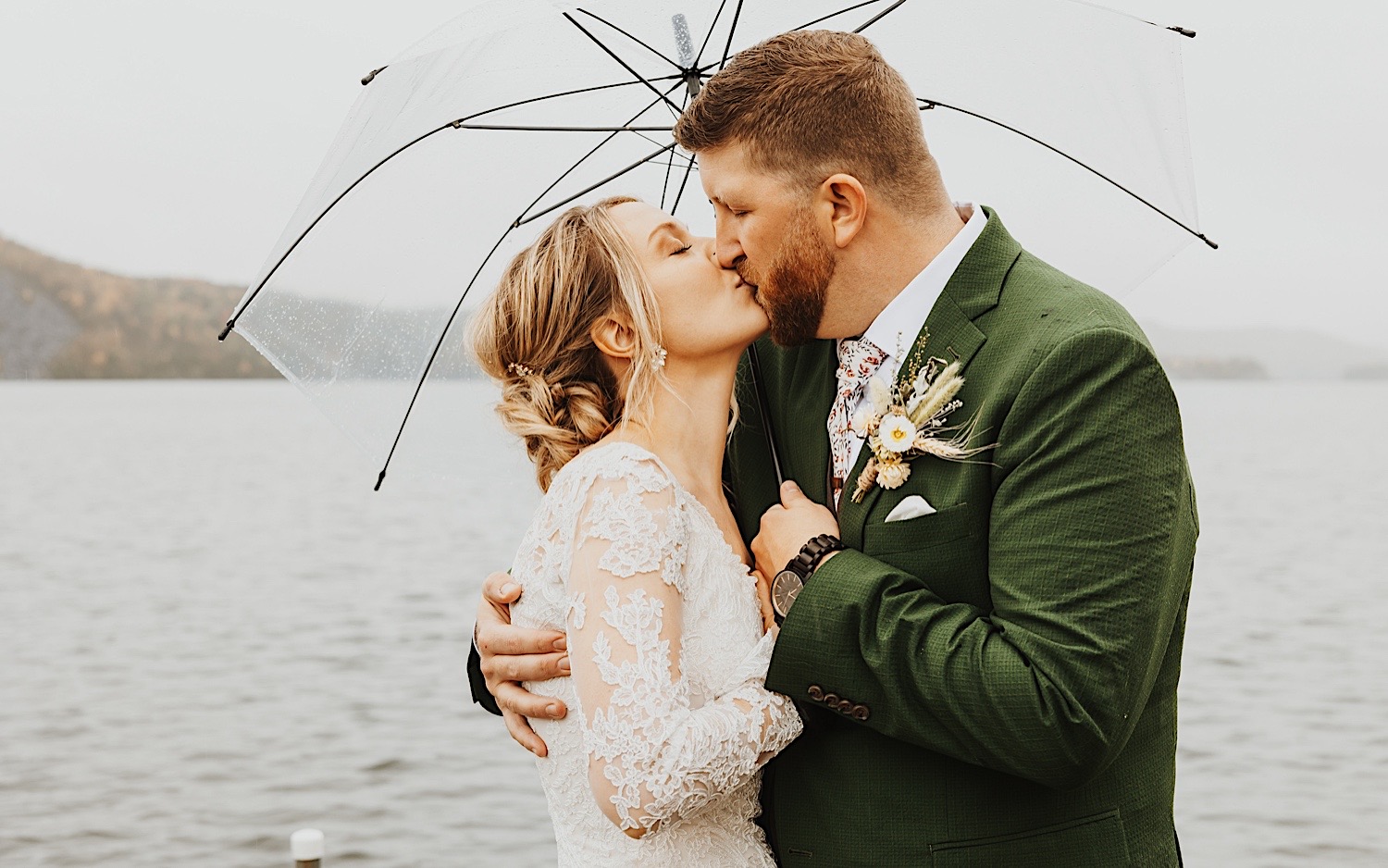A bride and groom kiss one another while under an umbrella as they stand on a dock of Lake Bomoseen in Vermont