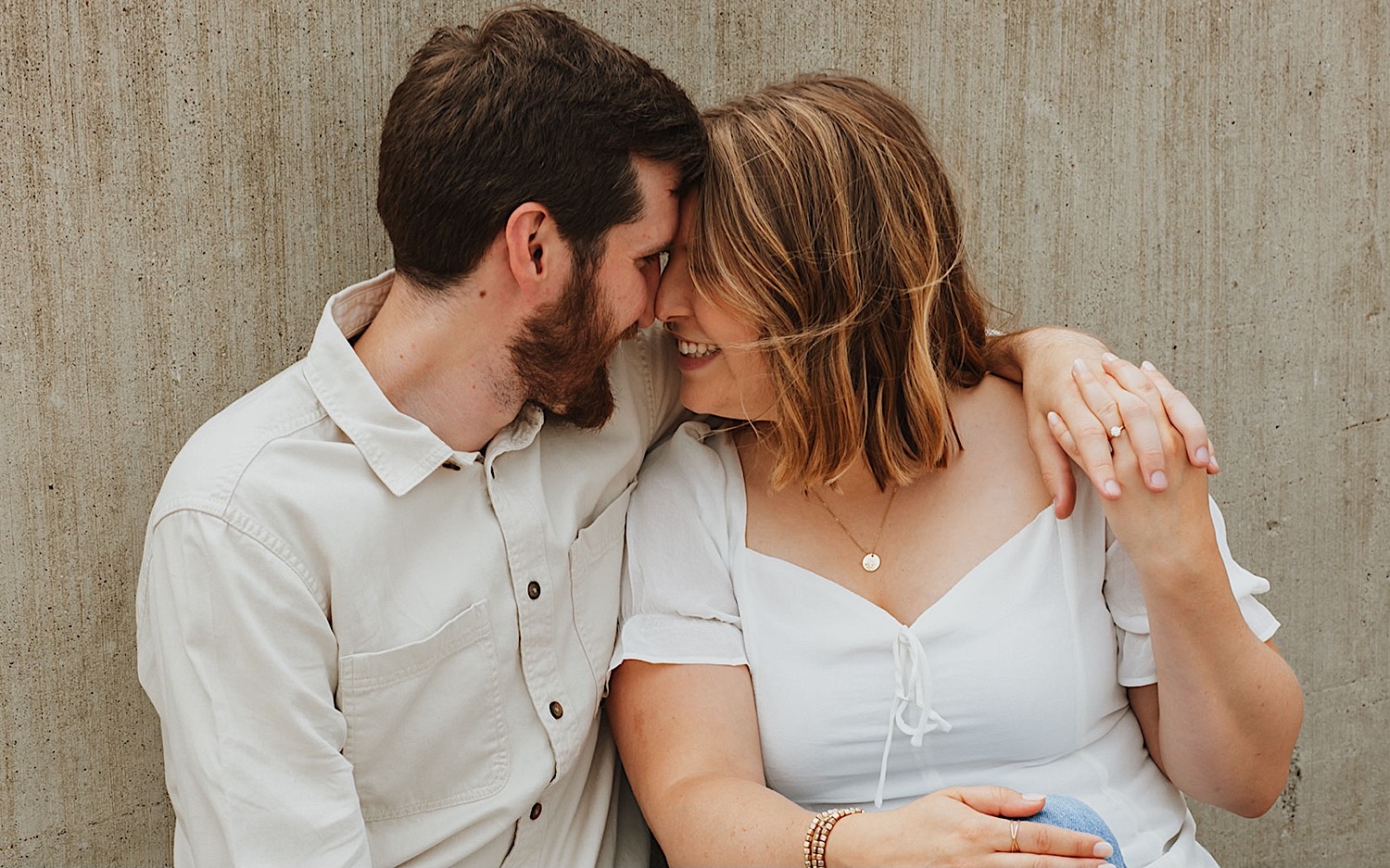 Close up photo of a couple smiling at one another as they sit side by side and lean in for a kiss