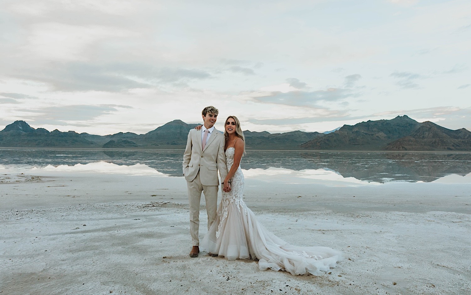 A bride and groom stand next to one another and hold hands while smiling at the camera standing in the Utah Salt Flats on their elopement day with the mountains behind them