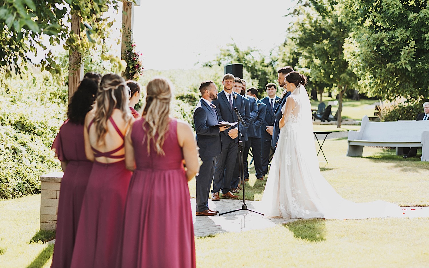 A bride and groom stand during their outdoor wedding ceremony at Legacy Hill Farm while their wedding parties watch on either side of them