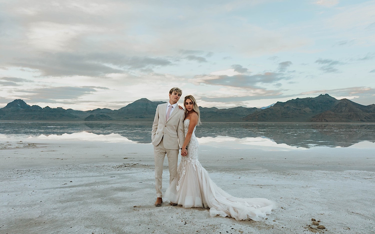A bride and groom stand next to one another and hold hands standing in the Utah Salt Flats on their elopement day with the mountains behind them
