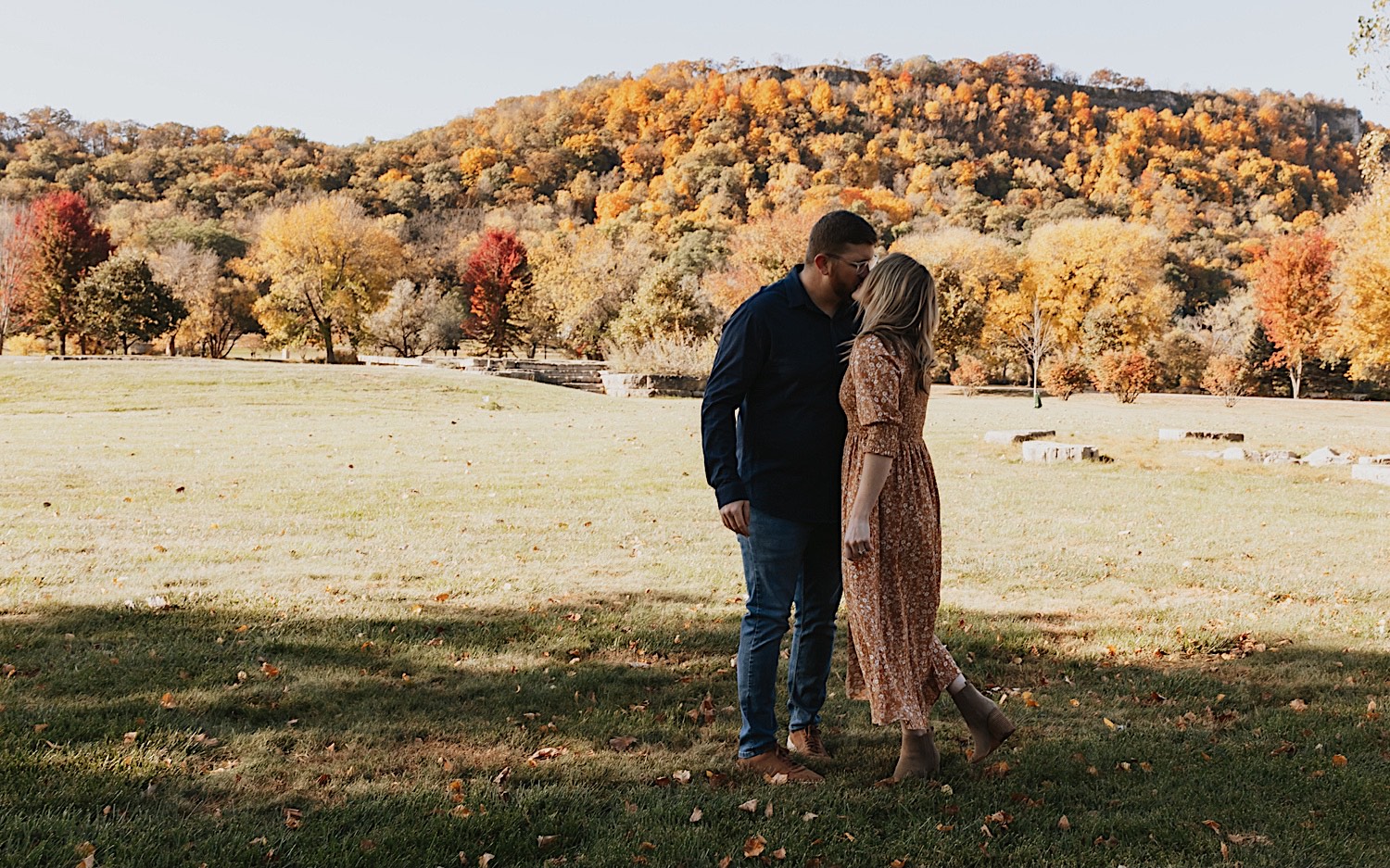 During a fall engagement session in Winona a couple kiss one another while standing in a park in front of a hillside where all the trees have changed colors