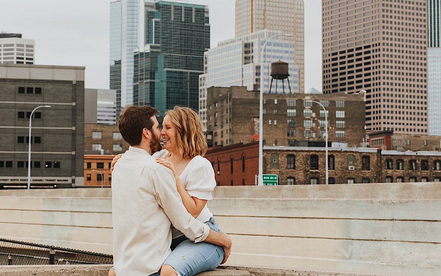 A woman sits on a ledge of a parking garage in downtown Minneapolis and smiles as a man hugs her and is about to kiss her while the two are having their engagement photos taken
