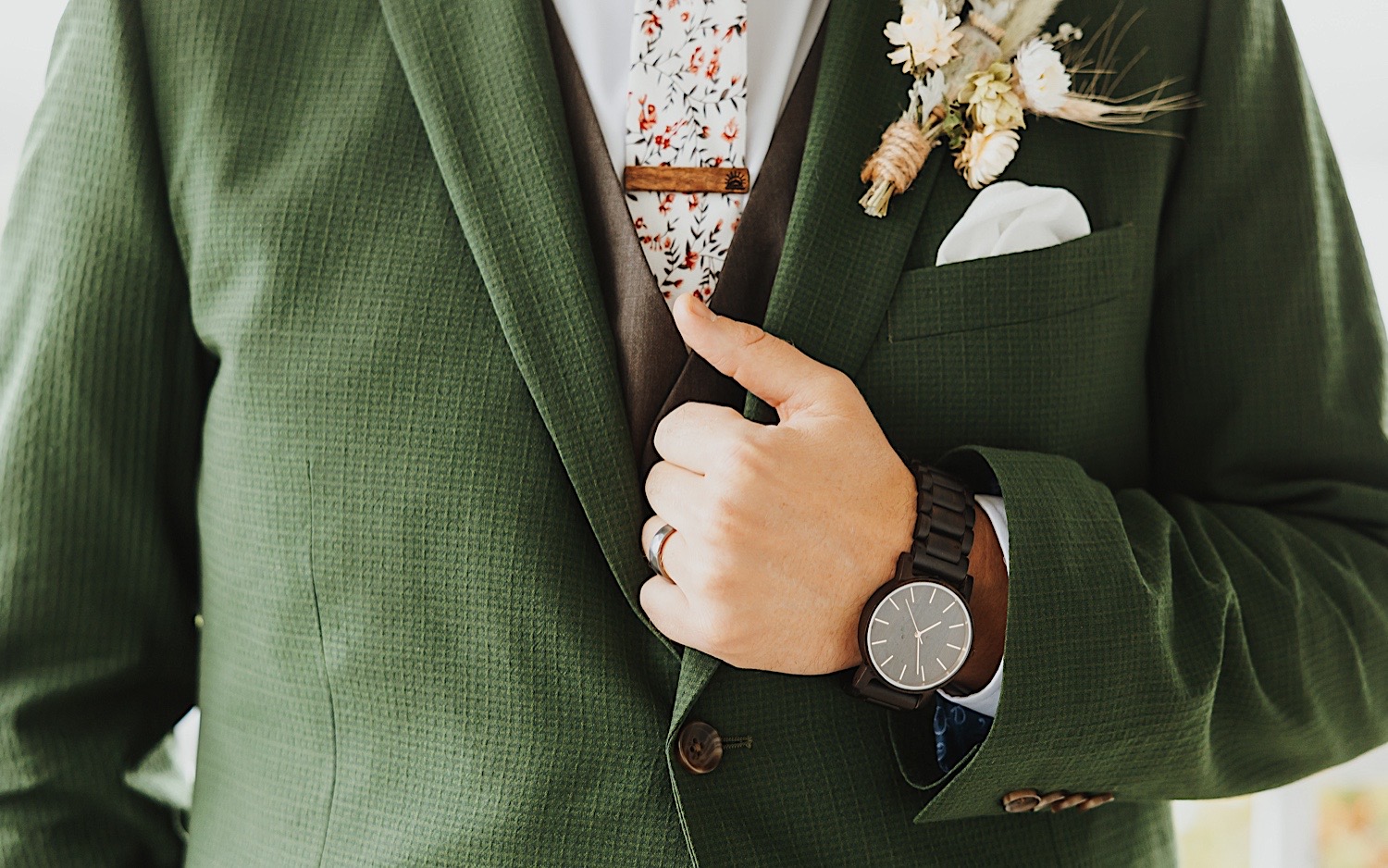 Close up photo of a groom's hand holding his green suit coat and showing off the watch on his wrist