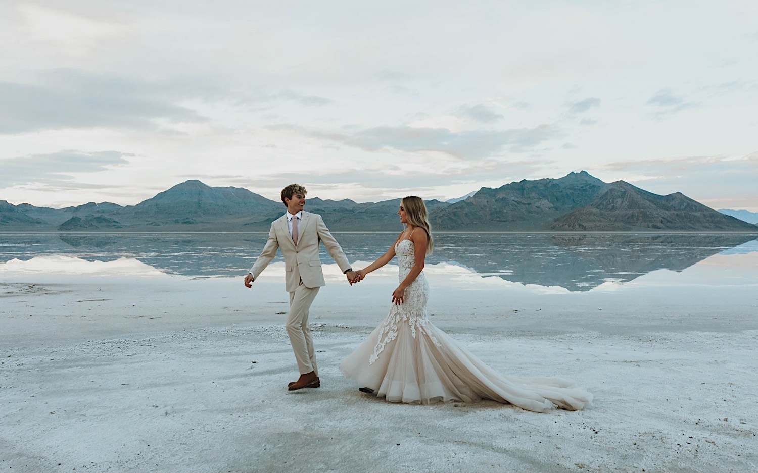In the Utah Salt Flats, a bride is led by her hand by a groom who smiles back at her during their elopement day with the mountains behind them