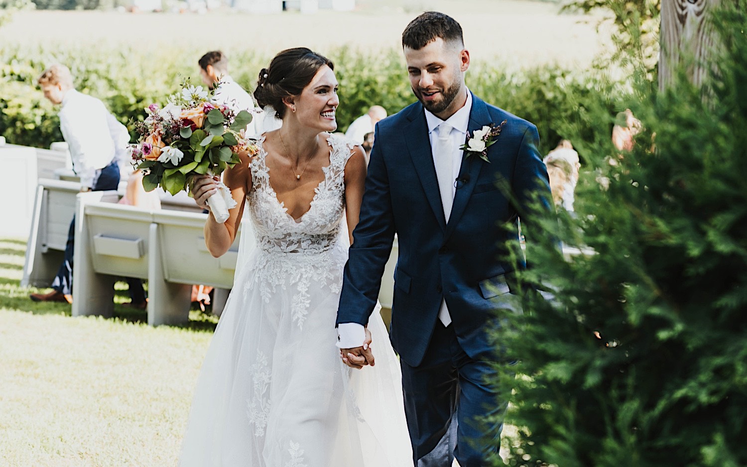 A bride smiles at the groom as the two walk away from their outdoor wedding ceremony at Legacy Hill Farm while holding hands