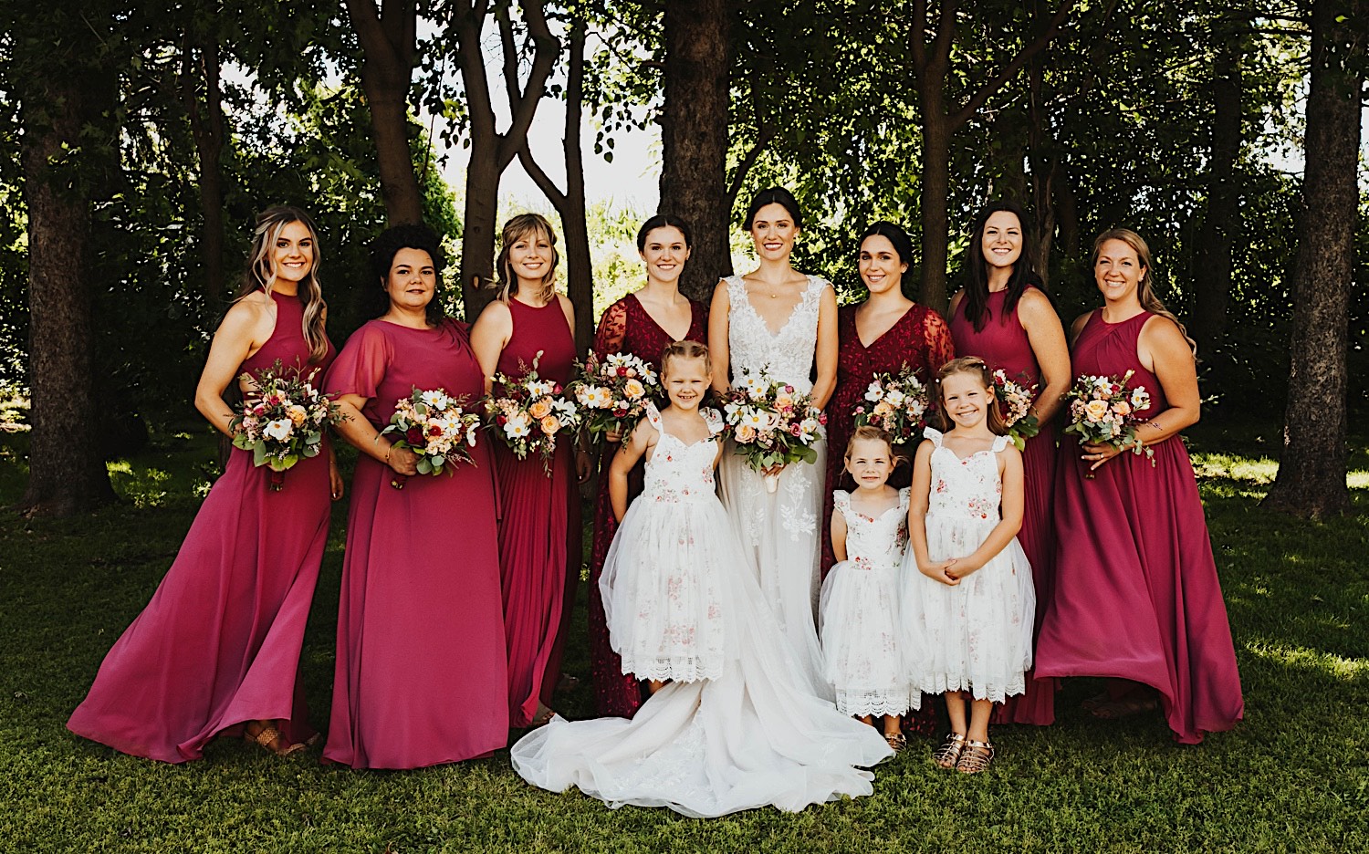 A bride stands in front of a row of trees and smiles with her bridesmaids and flower girls after a wedding ceremony at Legacy Hill Farm