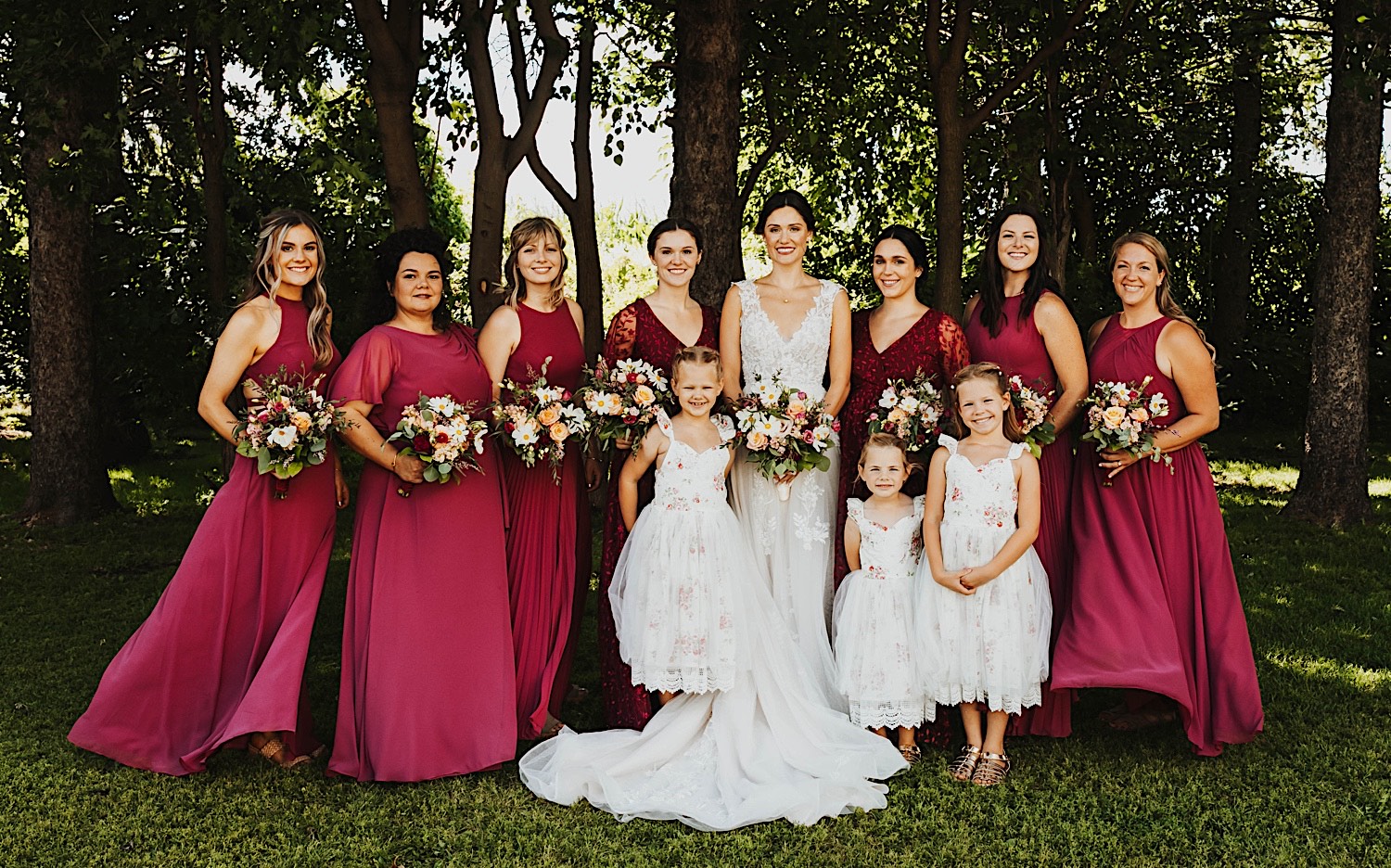 A bride stands in front of a row of trees and smiles with her bridesmaids and flower girls after a wedding ceremony at Legacy Hill Farm A bride stands in front of a row of trees and smiles with her bridesmaids and flower girls after a wedding ceremony at Legacy Hill Farm