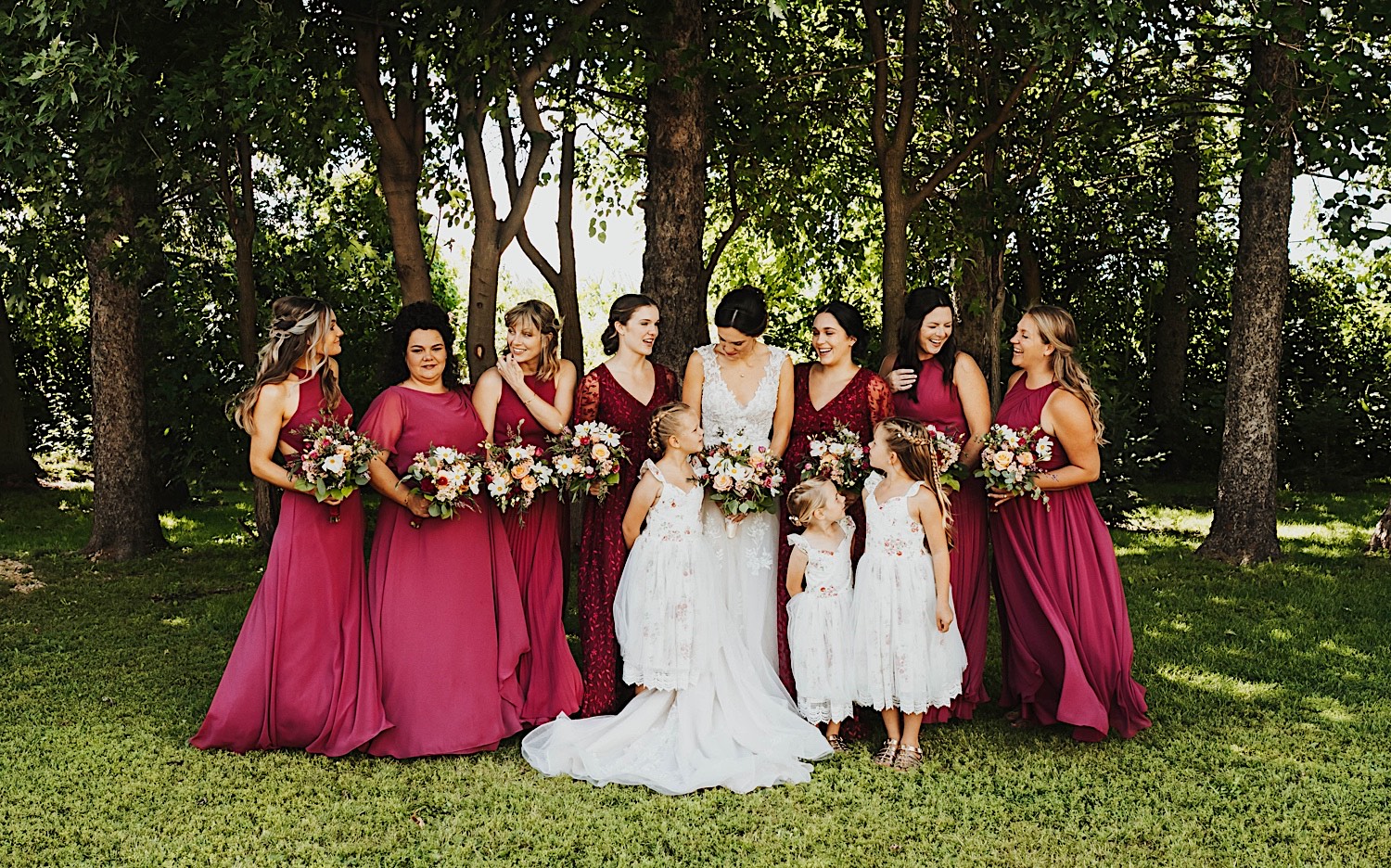 A bride mingles with her bridesmaids and flower girls while standing outside in front of a row of trees at the wedding venue Legacy Hill Farm