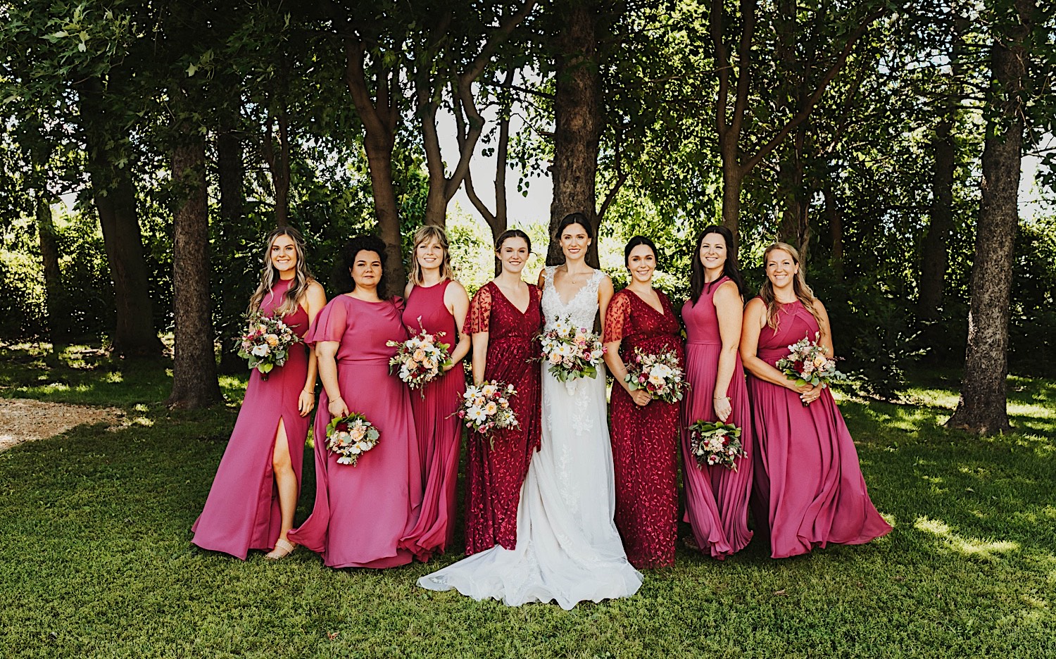 A bride smiles as she stands with her wedding party members in front of a row of trees at the wedding venue Legacy Hill Farm