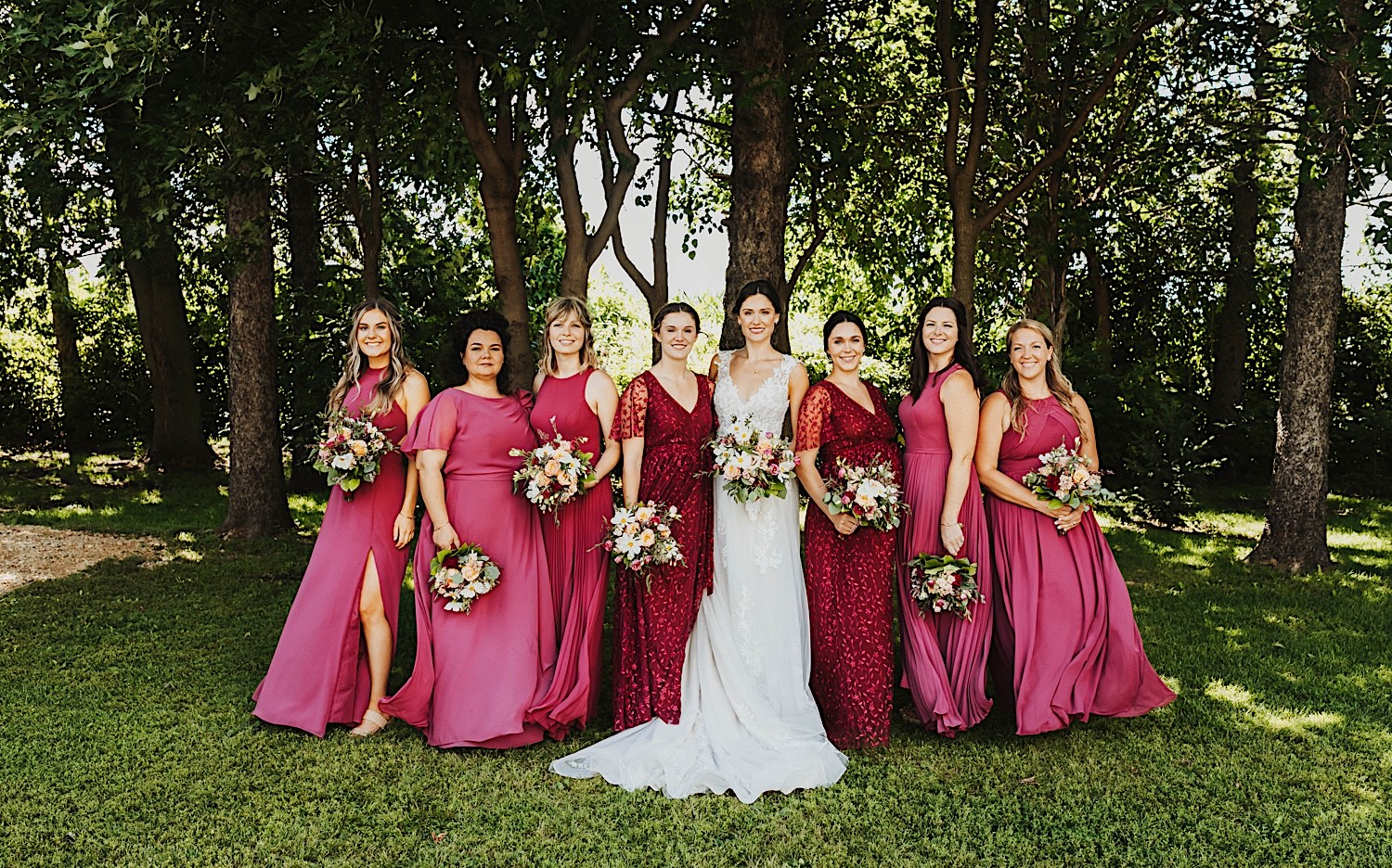 A bride smiles as she stands with her wedding party members in front of a row of trees at the wedding venue Legacy Hill Farm A bride smiles as she stands with her wedding party members in front of a row of trees at the wedding venue Legacy Hill Farm